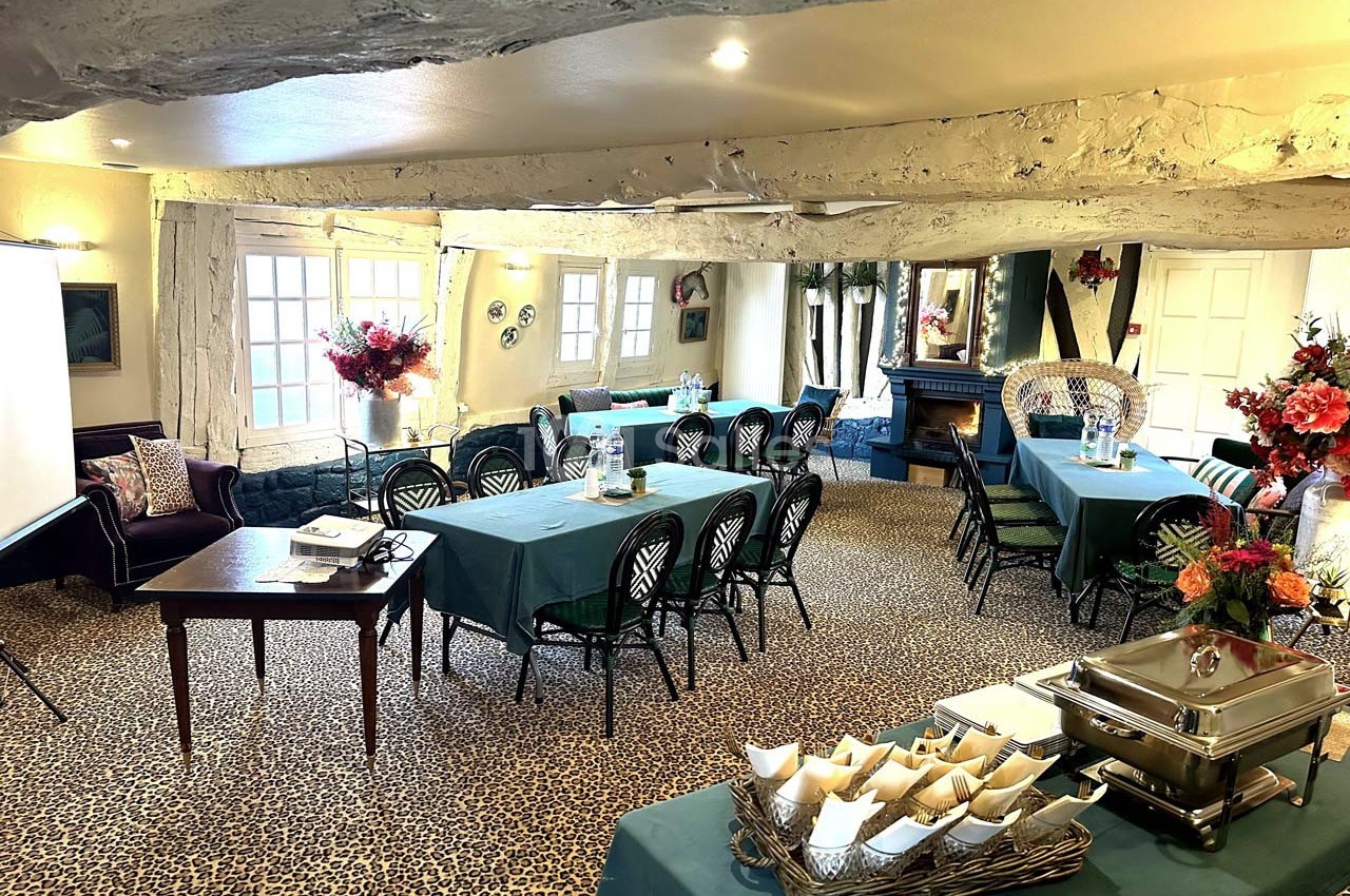 Salle de réunion lumineuse avec tables dressées, chaises noires, cheminée et décorations florales.