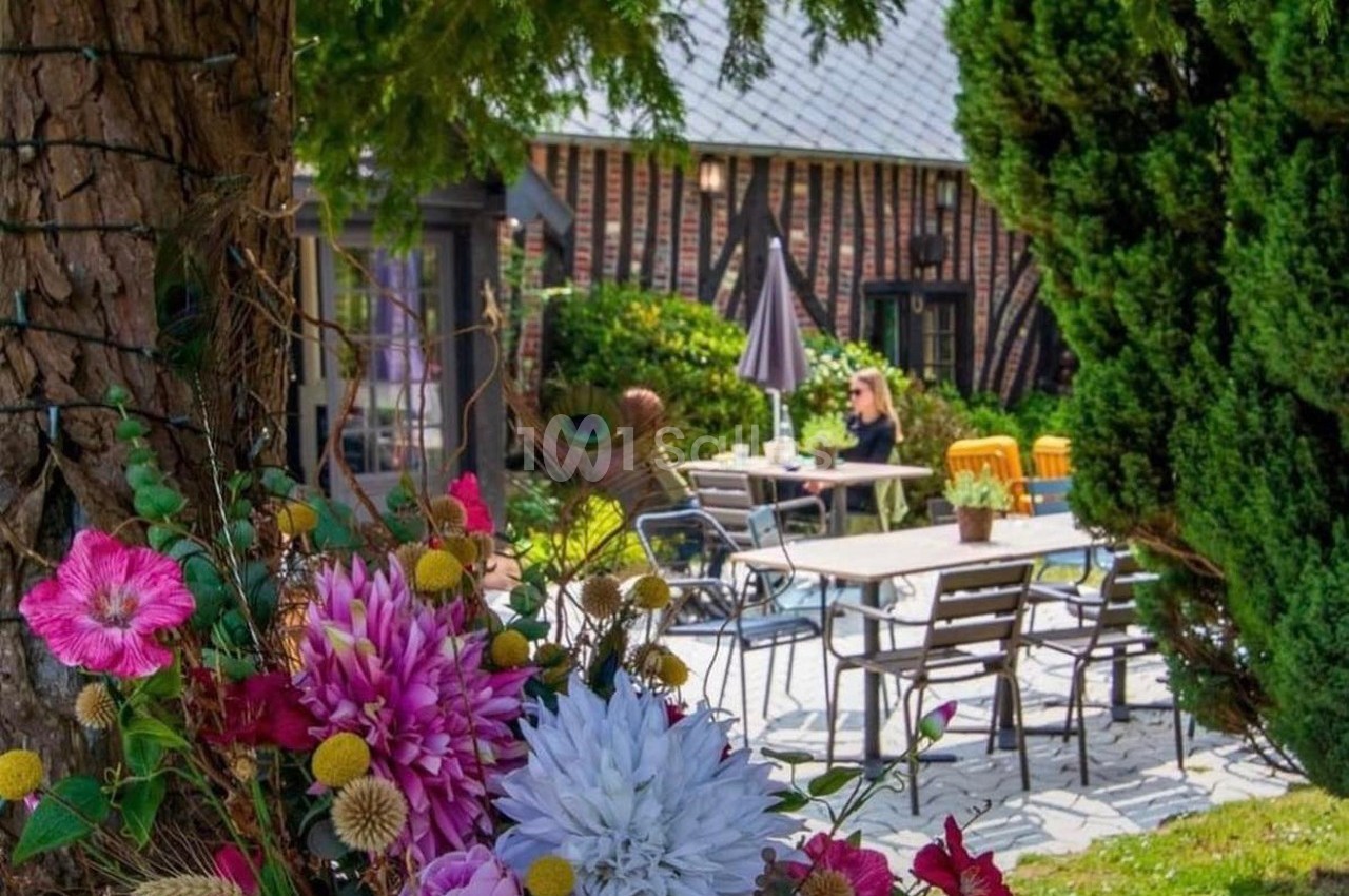 Terrasse extérieure avec tables et chaises, entourée de fleurs colorées et d'arbres, sous un ciel dégagé.