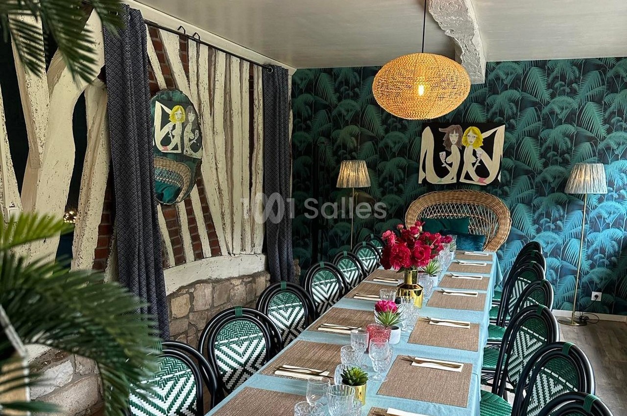 Salle à manger décorée avec des chaises en osier, une table dressée, un mur à motifs végétaux et des luminaires suspendus.