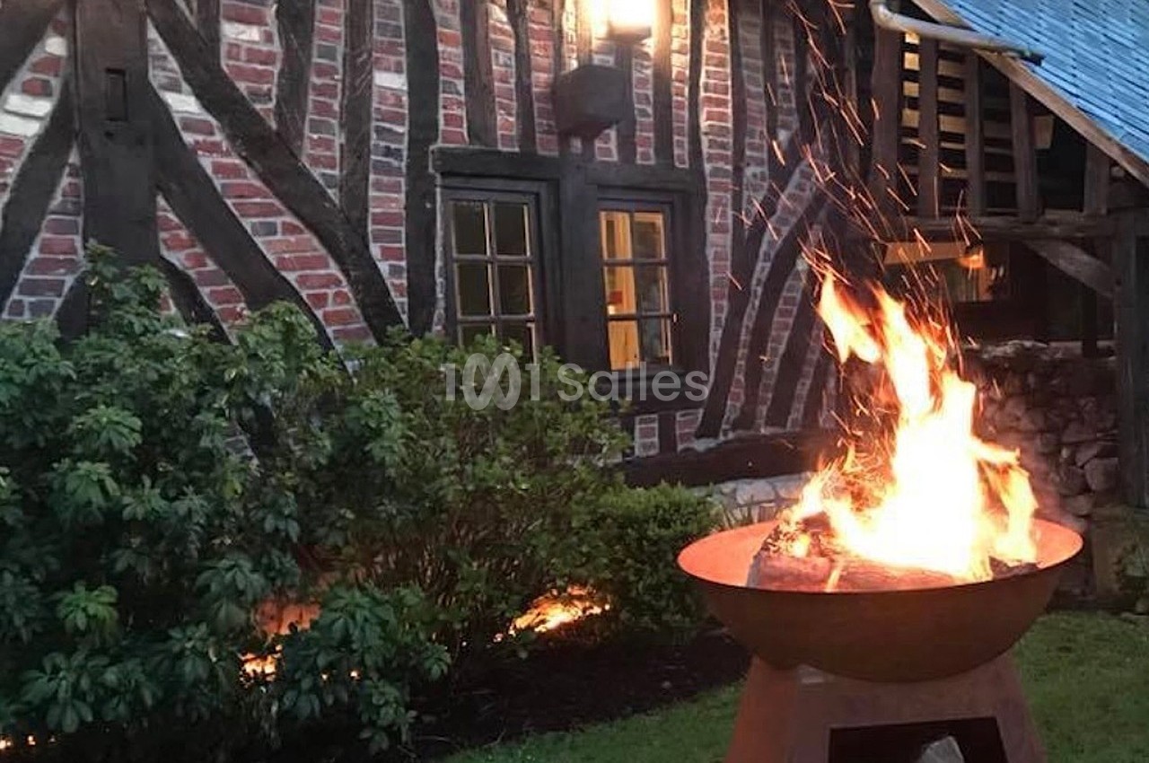 Brasero allumé devant une maison à colombages en briques et bois, entourée de végétation en soirée.