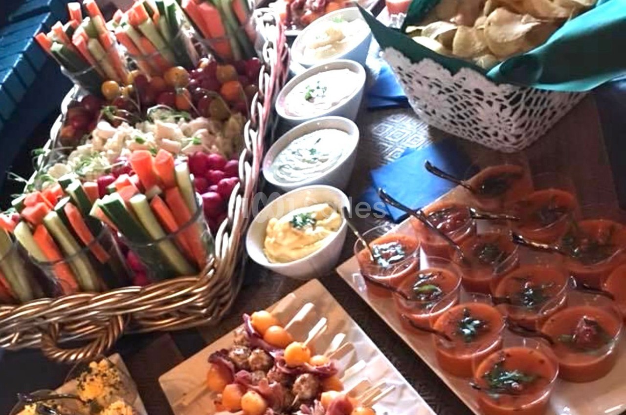 Plateau de buffet avec légumes frais, sauces, chips, brochettes et verrines de soupe disposés sur une table en bois.