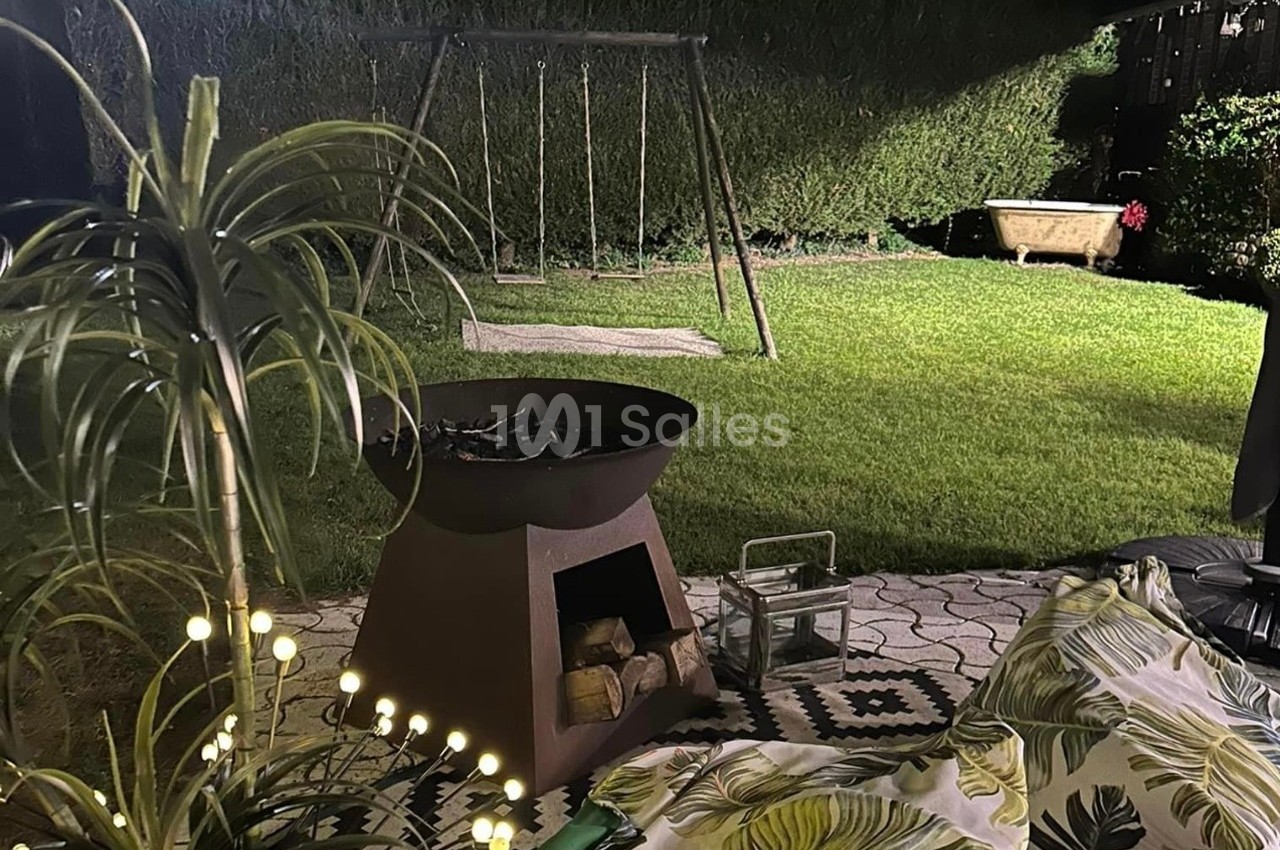 Brasero allumé sur une terrasse éclairée, avec vue sur un jardin, une balançoire et une baignoire ancienne en arrière-plan.