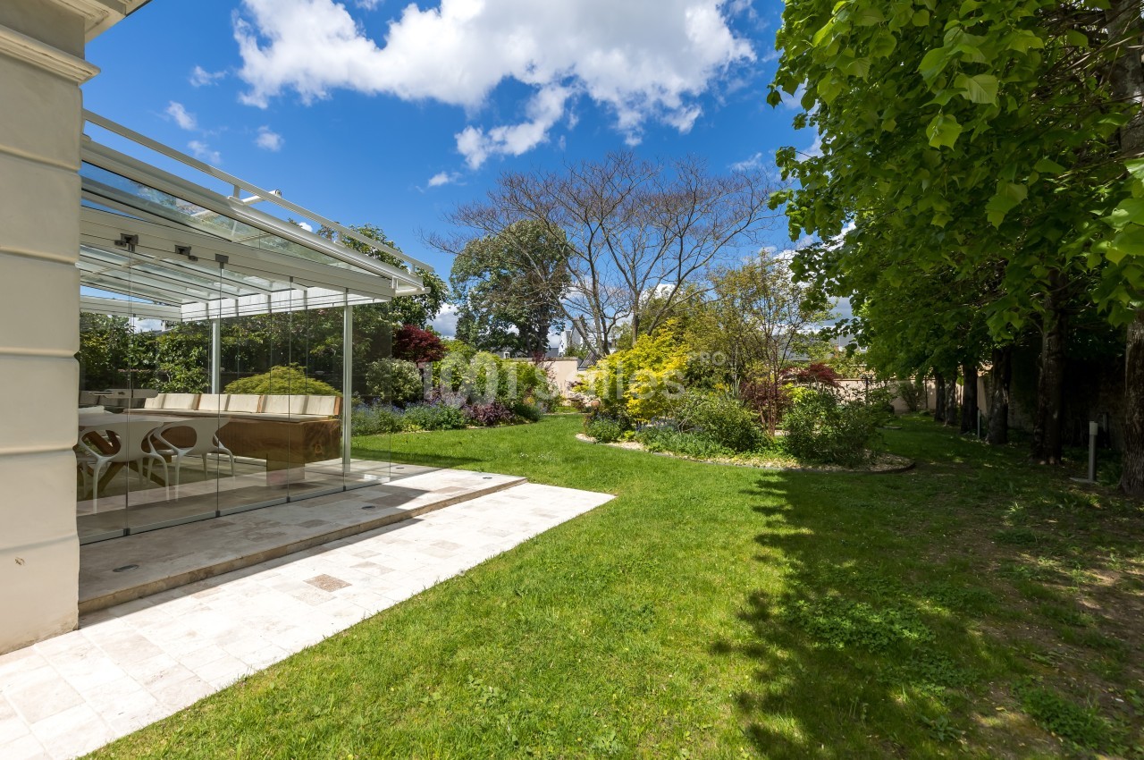 Jardin verdoyant avec pelouse, arbres et arbustes, jouxtant une véranda lumineuse sous un ciel partiellement nuageux.