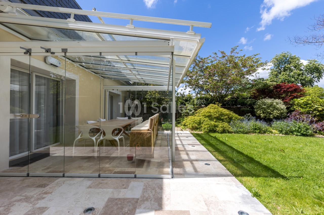 Véranda vitrée donnant sur une terrasse en pierre et un jardin verdoyant sous un ciel bleu.