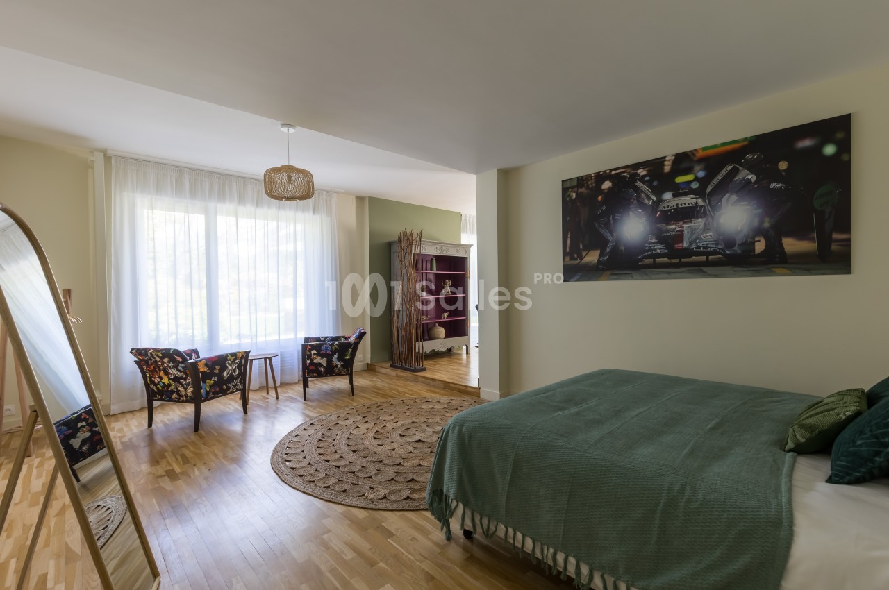 Chambre lumineuse avec lit double, coin salon, miroir sur pied, étagère et grande fenêtre avec rideaux blancs.