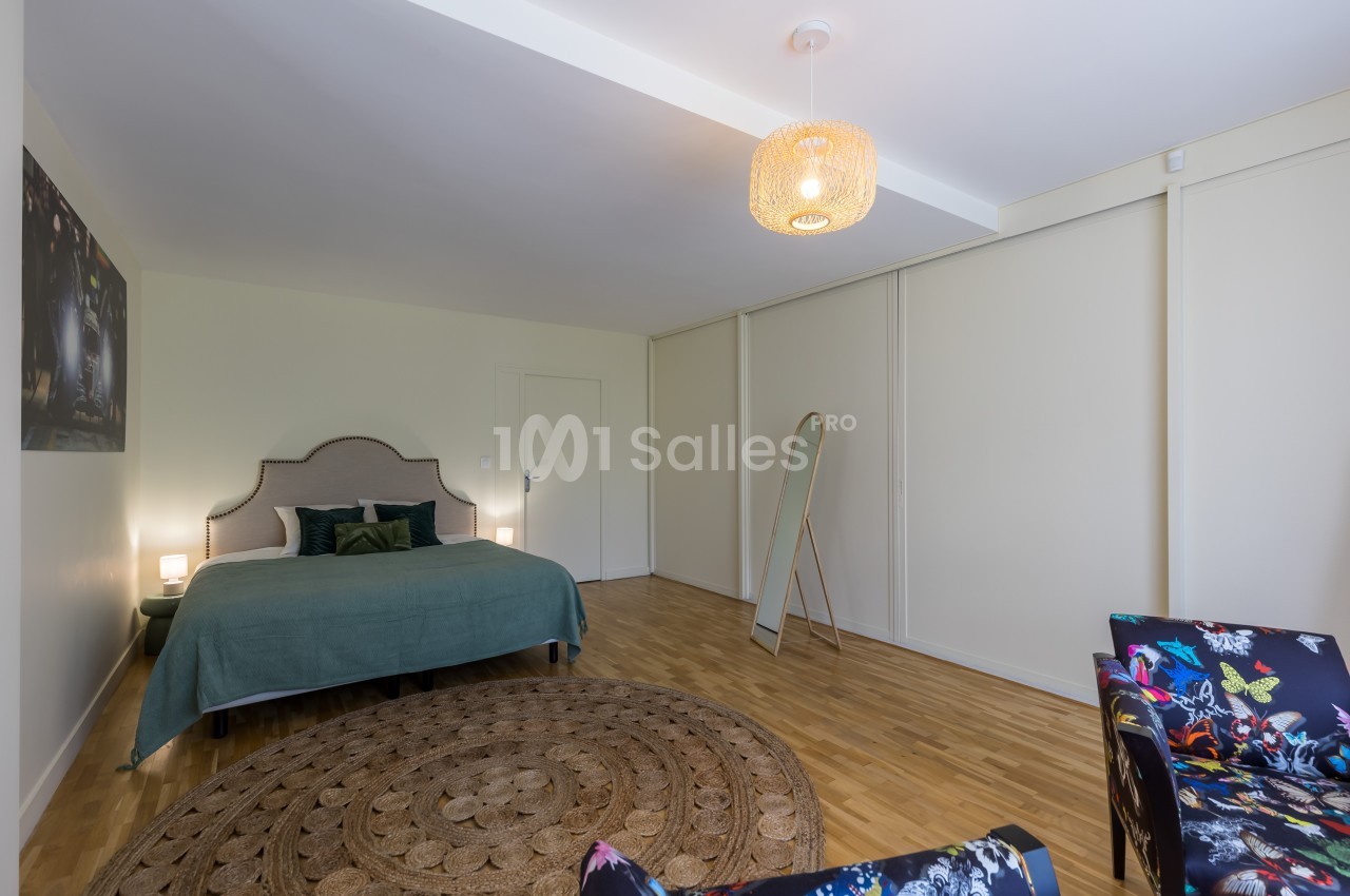 Chambre lumineuse avec lit double, tapis rond, miroir sur pied et fauteuil coloré, sur parquet en bois clair.