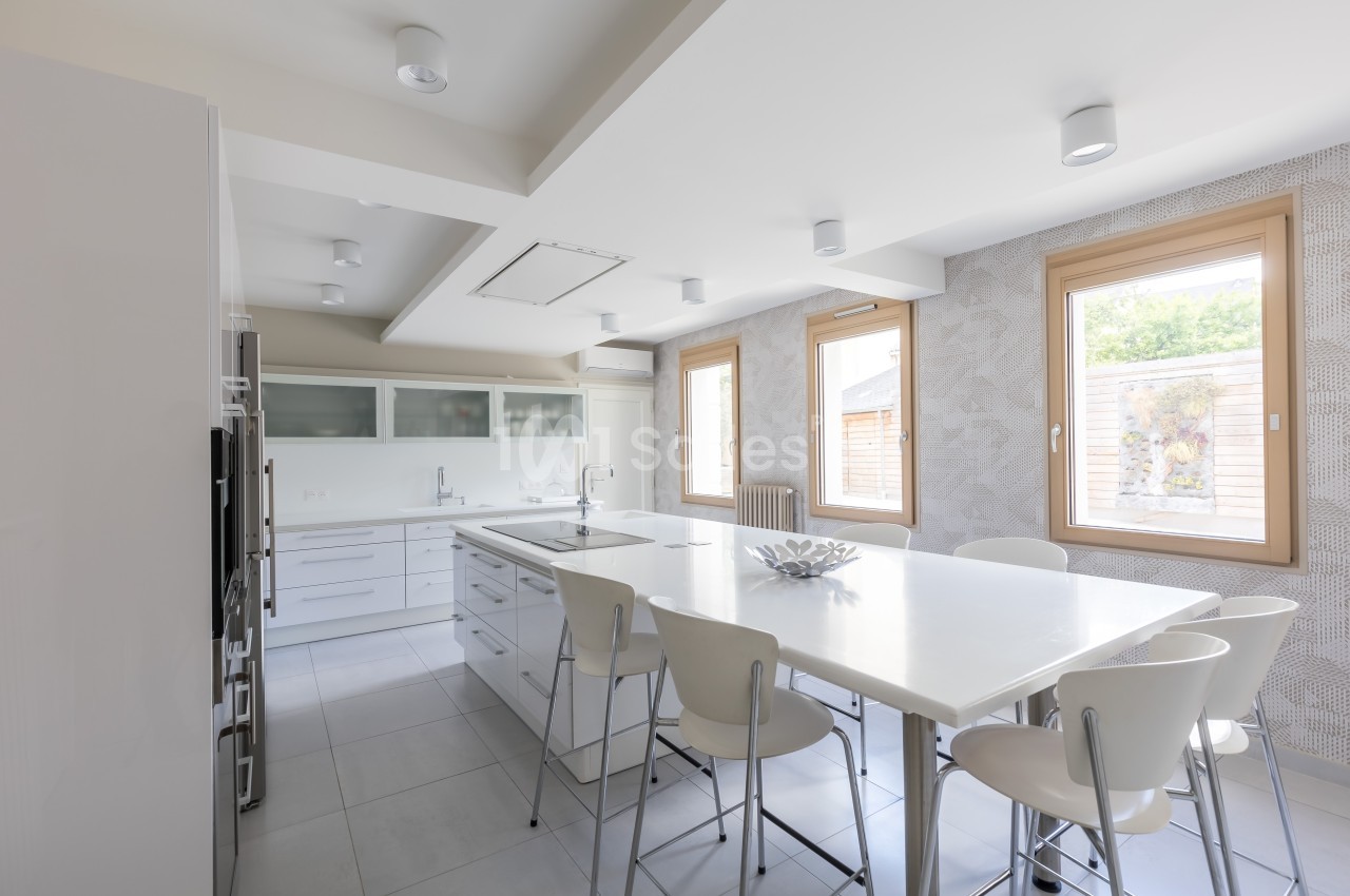 Cuisine moderne lumineuse avec un grand îlot central, des chaises blanches et des fenêtres laissant entrer la lumière…