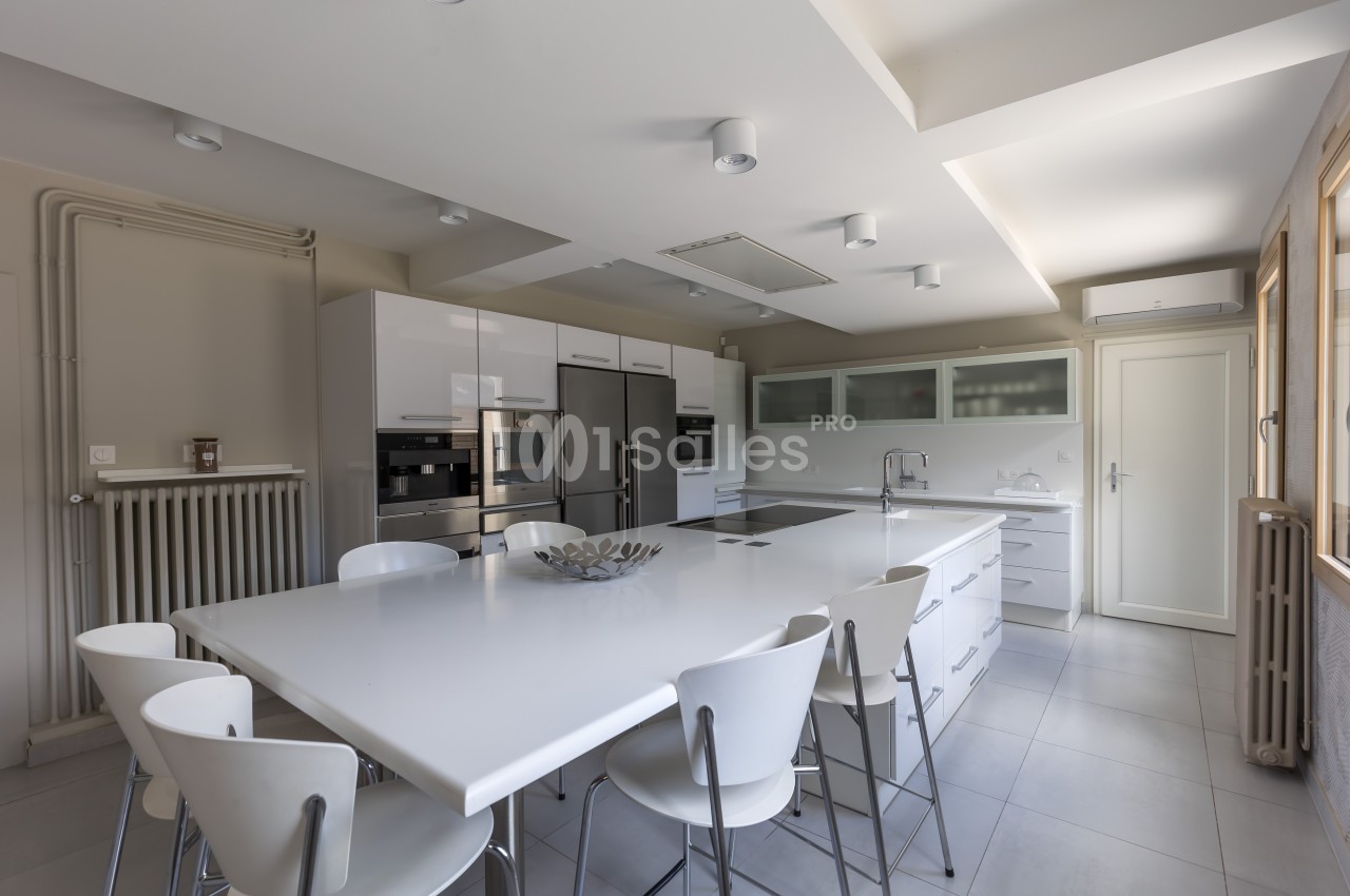 Cuisine moderne avec îlot central blanc, chaises assorties, électroménagers intégrés et grandes fenêtres laissant entrer la…