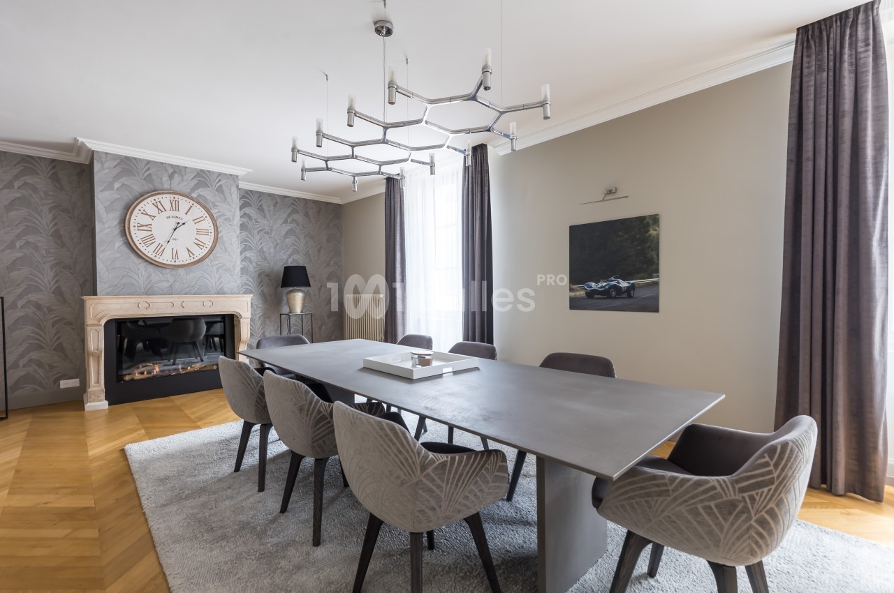 Salle à manger moderne avec table en métal, chaises grises, cheminée, horloge murale et tableau décoratif.