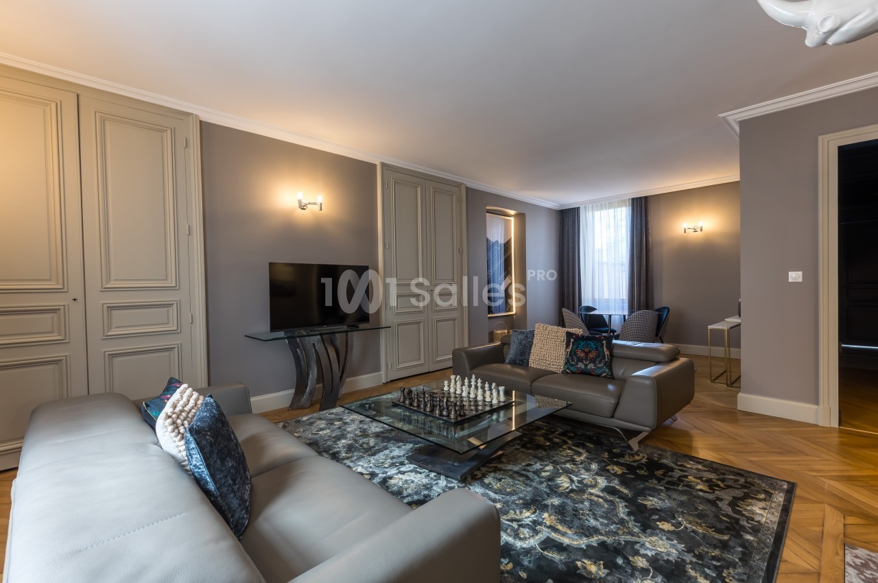 Salon élégant avec canapés gris, table basse avec échiquier, tapis décoratif et éclairage mural doux.