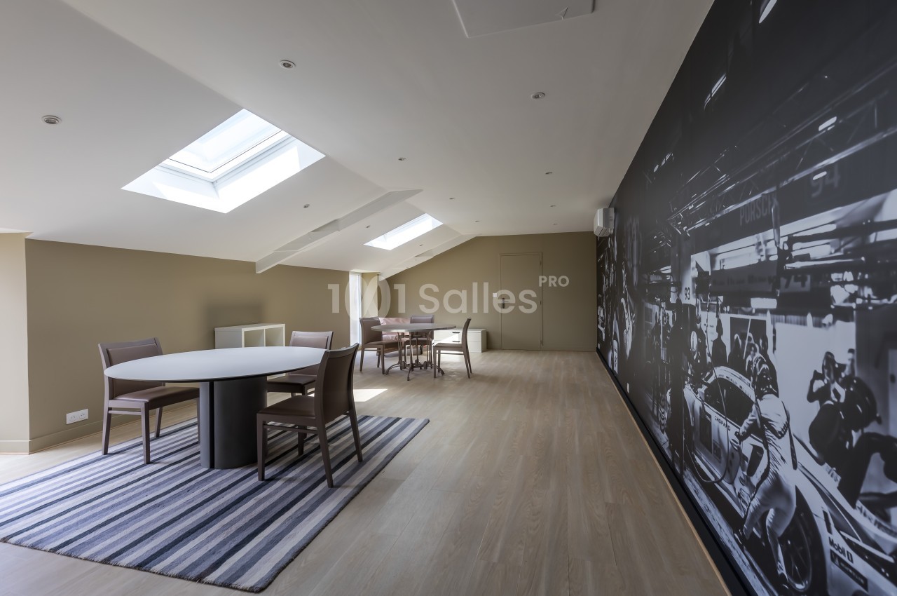 Salle lumineuse sous combles avec table ronde, chaises, tapis rayé et mur décoré d'une fresque en noir et blanc.