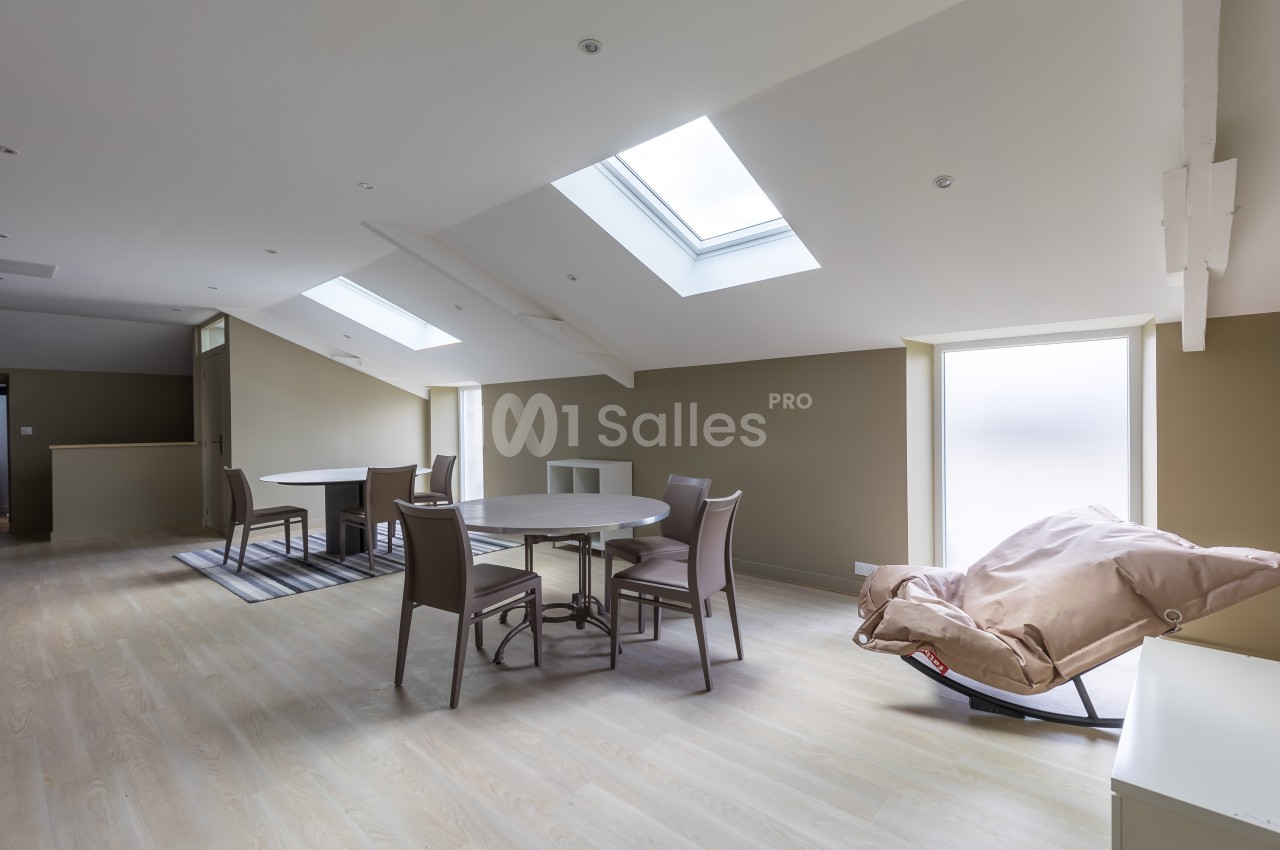 Salle à manger lumineuse sous combles avec table ronde, chaises, fauteuil inclinable et puits de lumière.