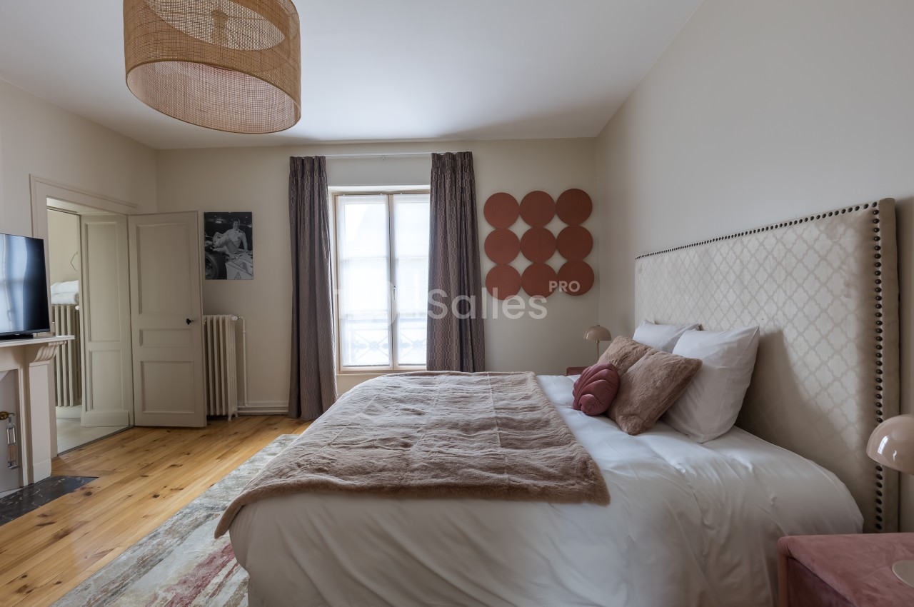 Chambre lumineuse avec un lit double, tête de lit capitonnée, décor minimaliste et fenêtre avec rideaux.