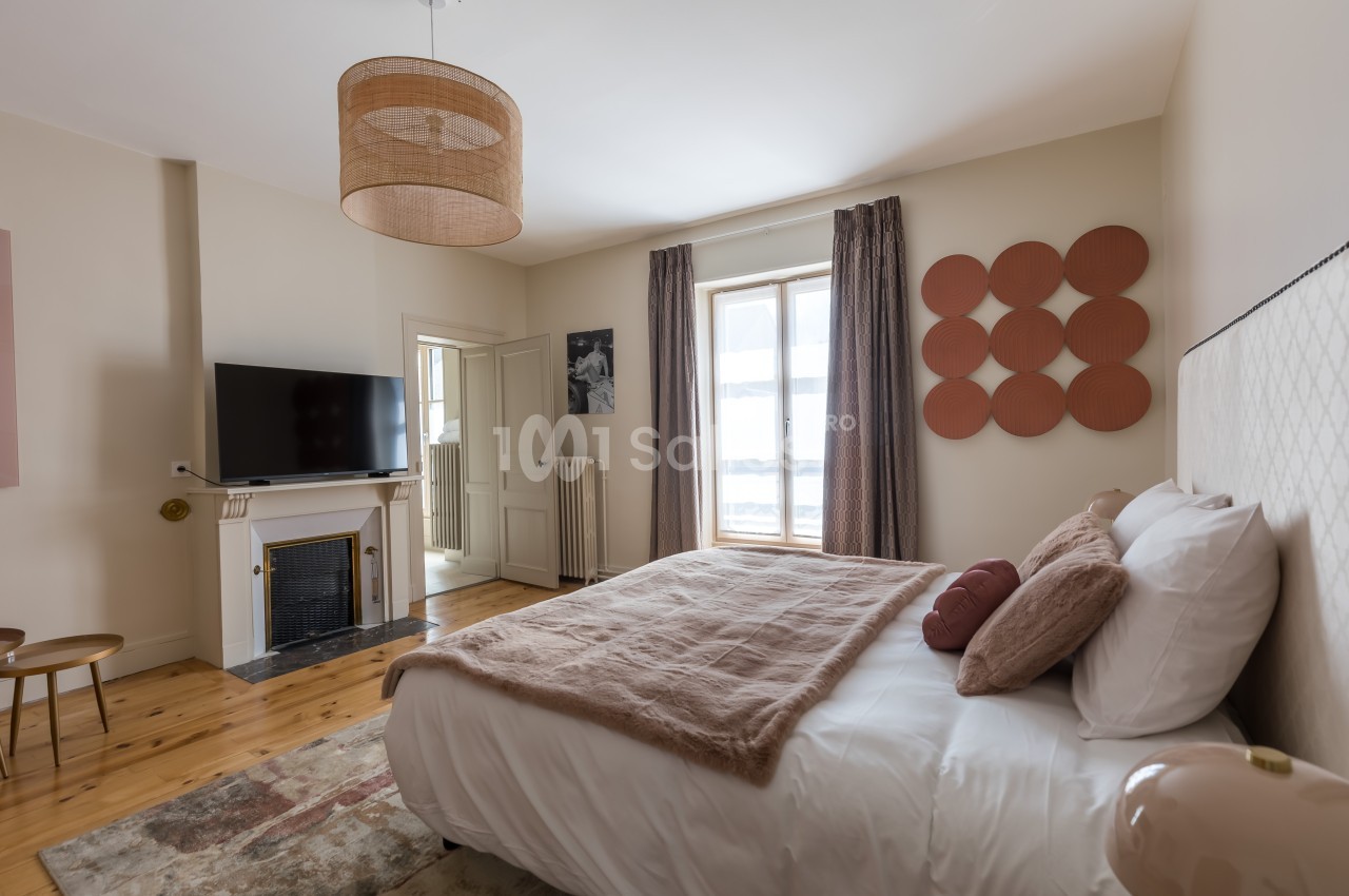 Chambre lumineuse avec lit double, télévision, cheminée, décoration murale et grandes fenêtres avec rideaux.
