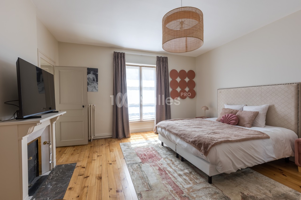 Chambre lumineuse avec lit double, parquet en bois, tapis, télévision, cheminée et grande fenêtre avec rideaux.