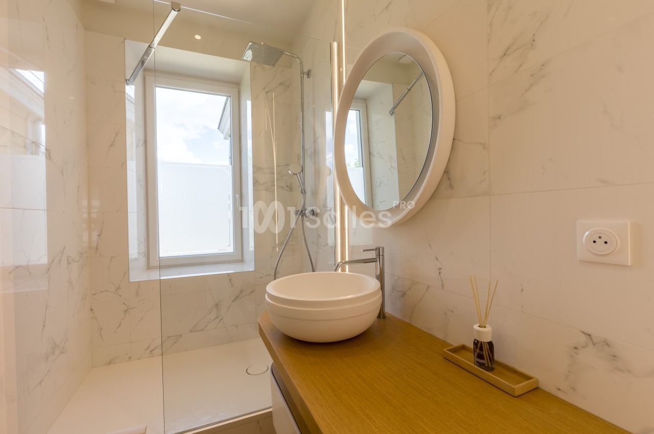 Salle de bain moderne avec lavabo rond, miroir éclairé, douche vitrée et murs en carrelage effet marbre.
