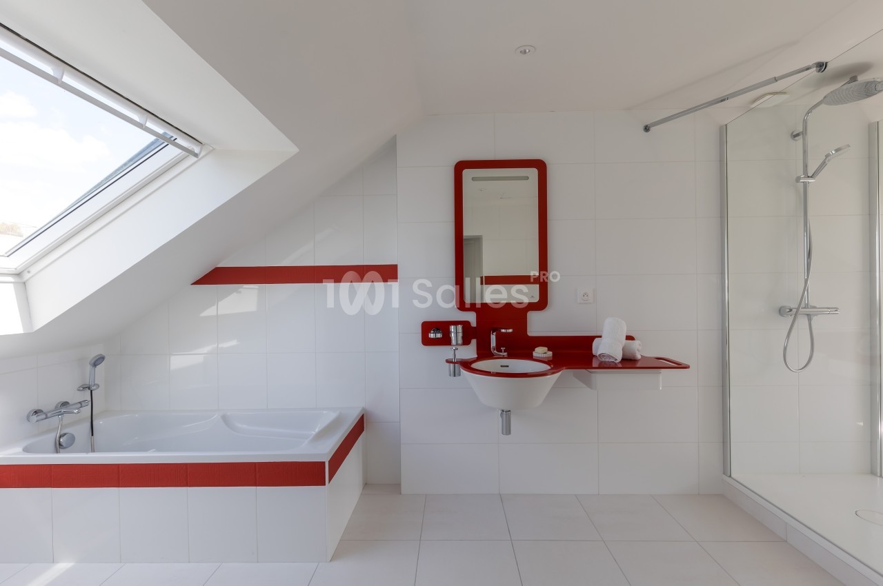 Salle de bain moderne avec baignoire, douche, lavabo et miroir encadrés de rouge, sous un toit mansardé avec fenêtre.