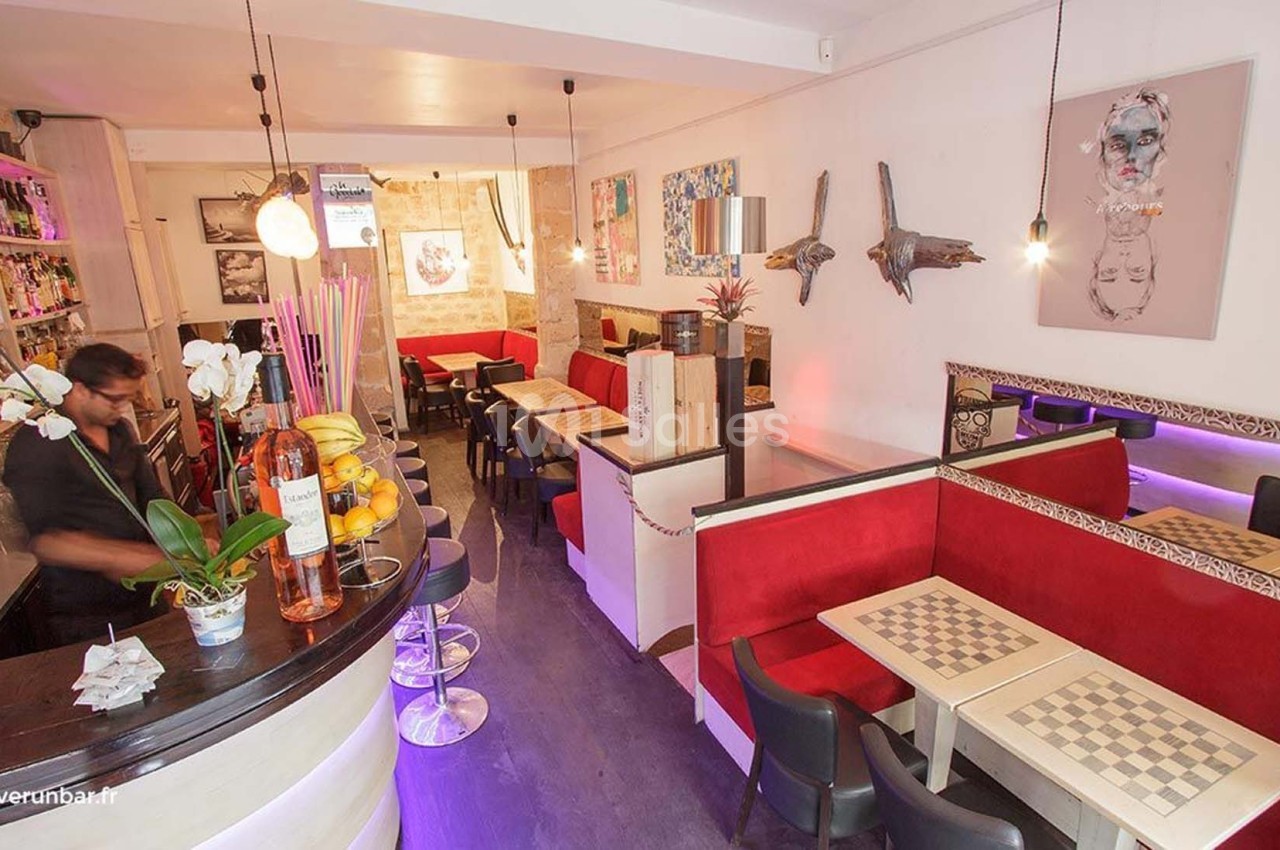Intérieur d'un bar avec comptoir, tables à damier, banquettes rouges et décorations murales artistiques.
