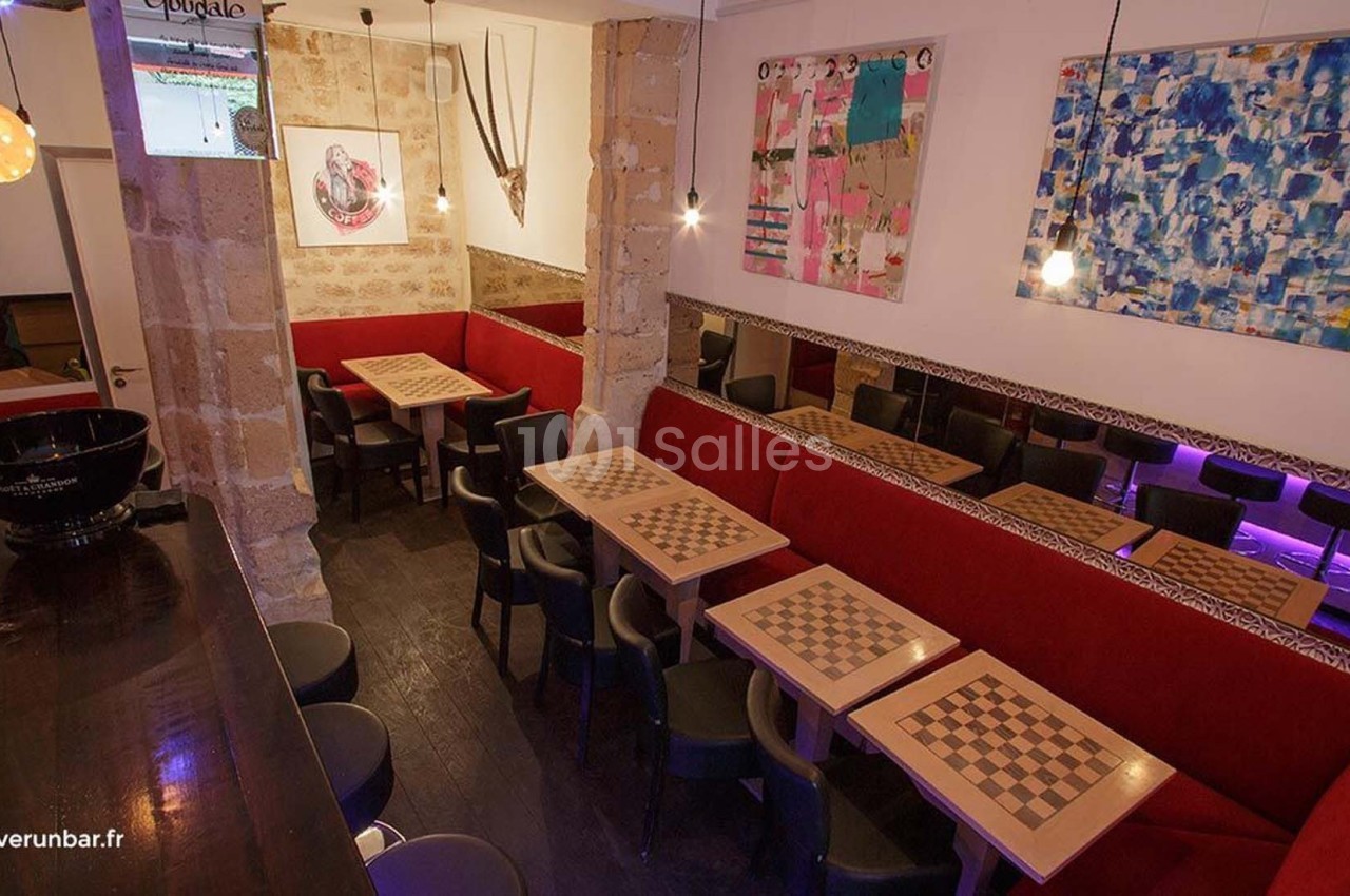 Salle de bar avec tables de jeu d'échecs, banquettes rouges, chaises noires et décorations murales colorées.