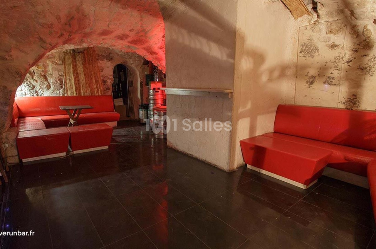 Salle voûtée avec murs en pierre, banquettes rouges, sol sombre et éclairage tamisé.