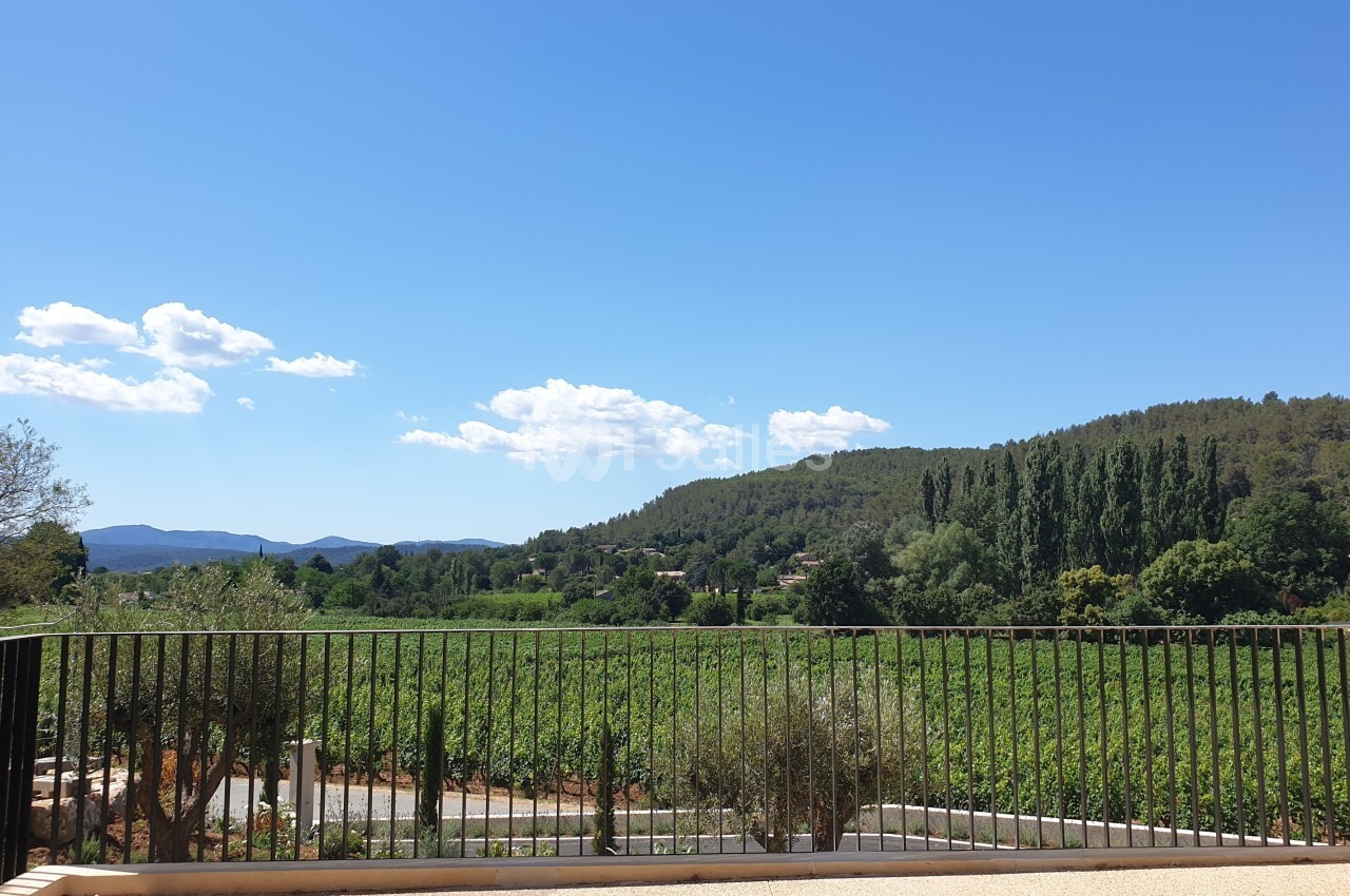 Vue sur un paysage rural avec des collines boisées, des vignes et un ciel dégagé, depuis une terrasse avec une barrière…