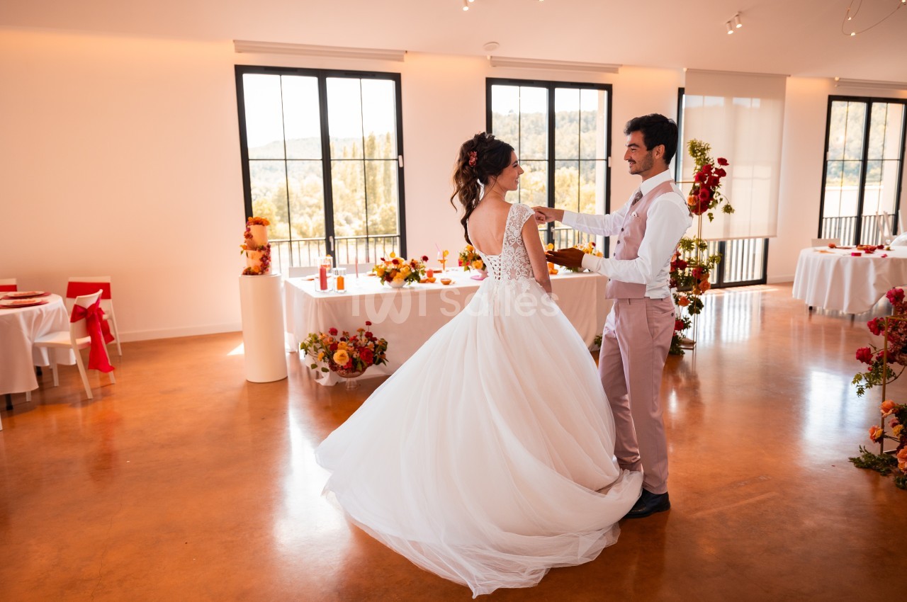 Un couple danse dans une salle lumineuse décorée de fleurs et de tables pour une réception.