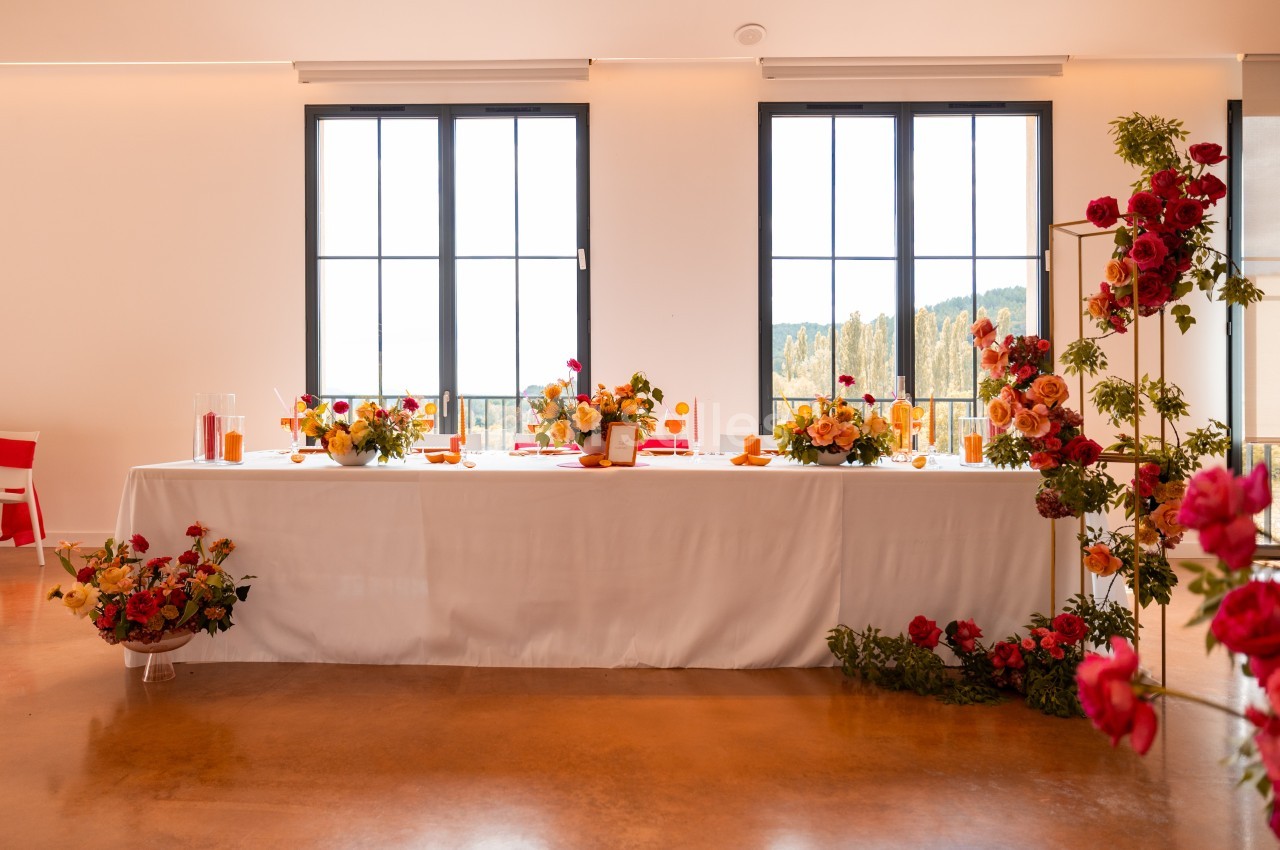 Table décorée avec des fleurs colorées, des bougies et des éléments de vaisselle, placée devant de grandes fenêtres.