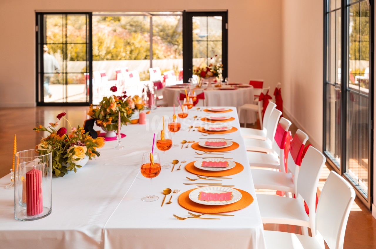 Table dressée avec des assiettes colorées, verres orange et décorations florales dans une salle lumineuse avec grandes baies…
