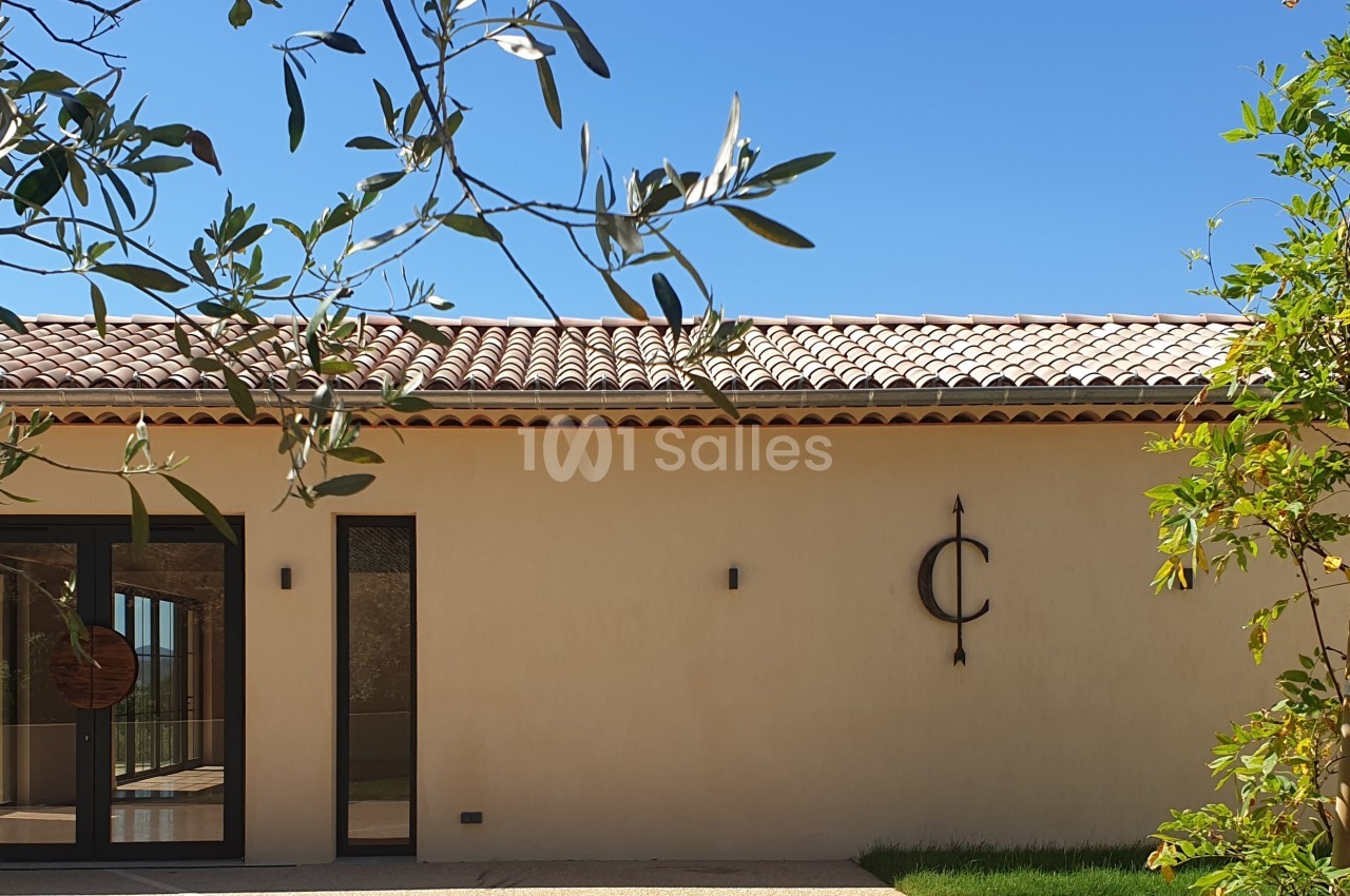 Façade d'une maison beige avec toit en tuiles, porte vitrée, et végétation au premier plan sous un ciel bleu.
