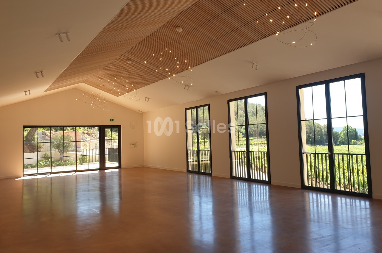 Salle lumineuse avec de grandes baies vitrées, un plafond en bois incliné et des lumières suspendues.