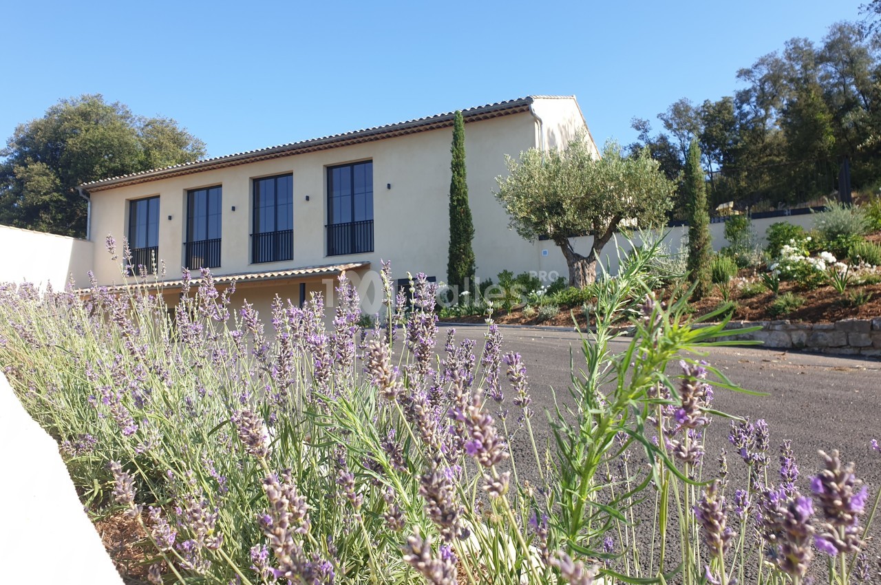 Façade d'une maison beige à toit en tuiles, entourée de lavandes et d'arbres, sous un ciel bleu.