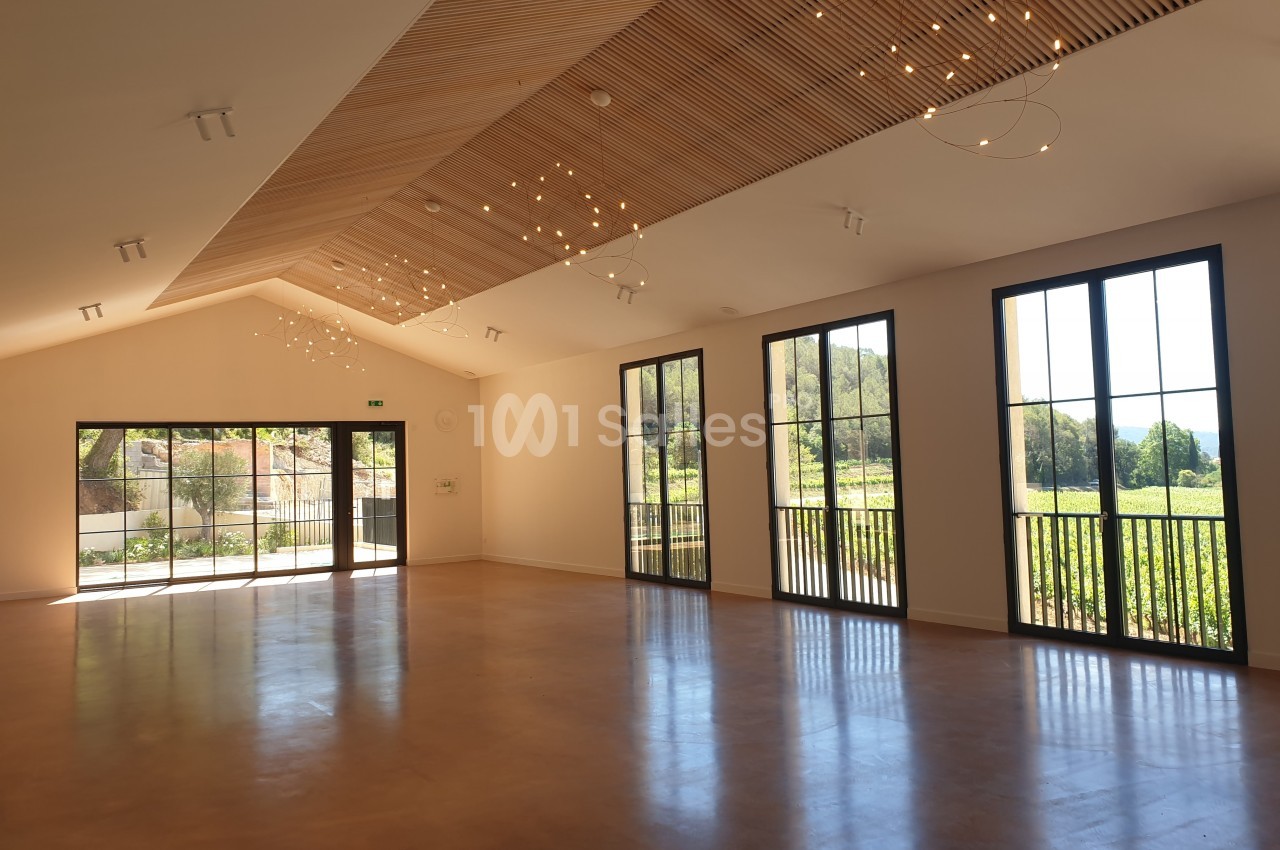 Salle lumineuse avec de grandes baies vitrées, un plafond en bois incliné et des luminaires suspendus.