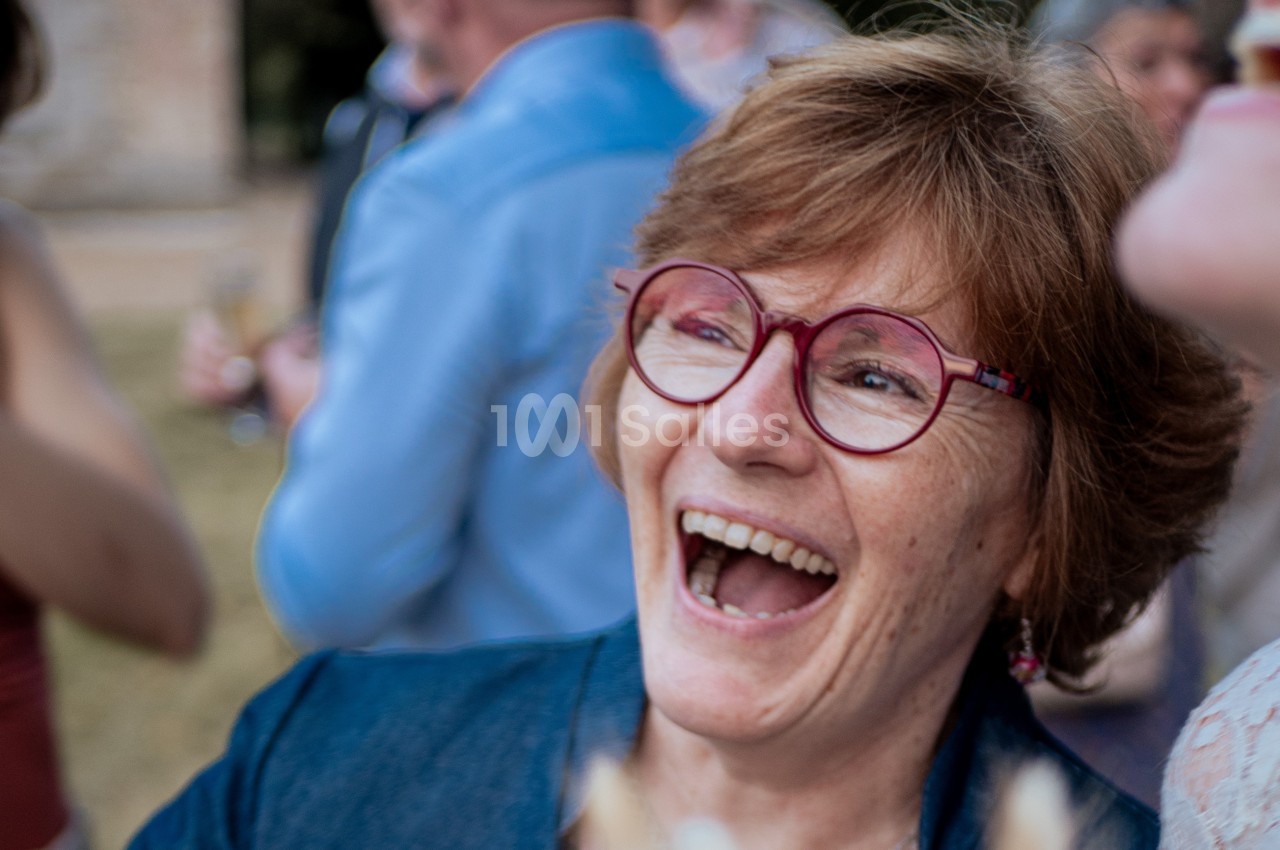 Femme souriante portant des lunettes rouges, entourée de personnes floues en arrière-plan lors d'un événement en extérieur.