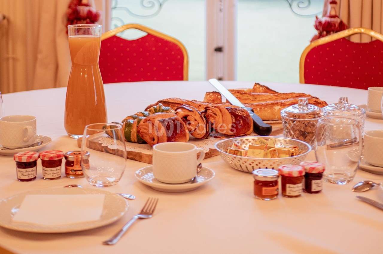 Petit-déjeuner servi sur une table avec viennoiseries, jus d'orange, confitures, tasses et couverts.