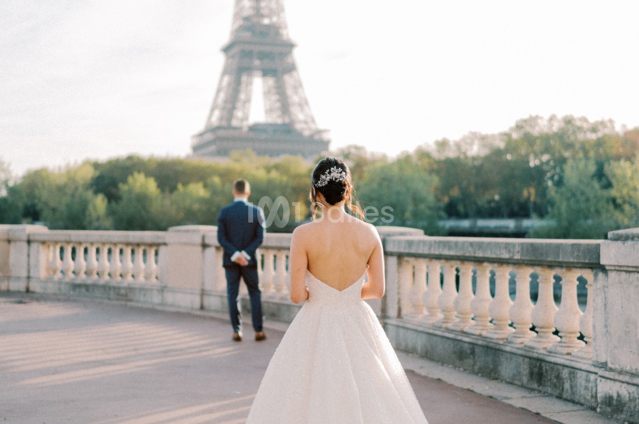 Une femme en robe de mariée marche sur un pont avec la tour Eiffel en arrière-plan, un homme lui tournant le dos.