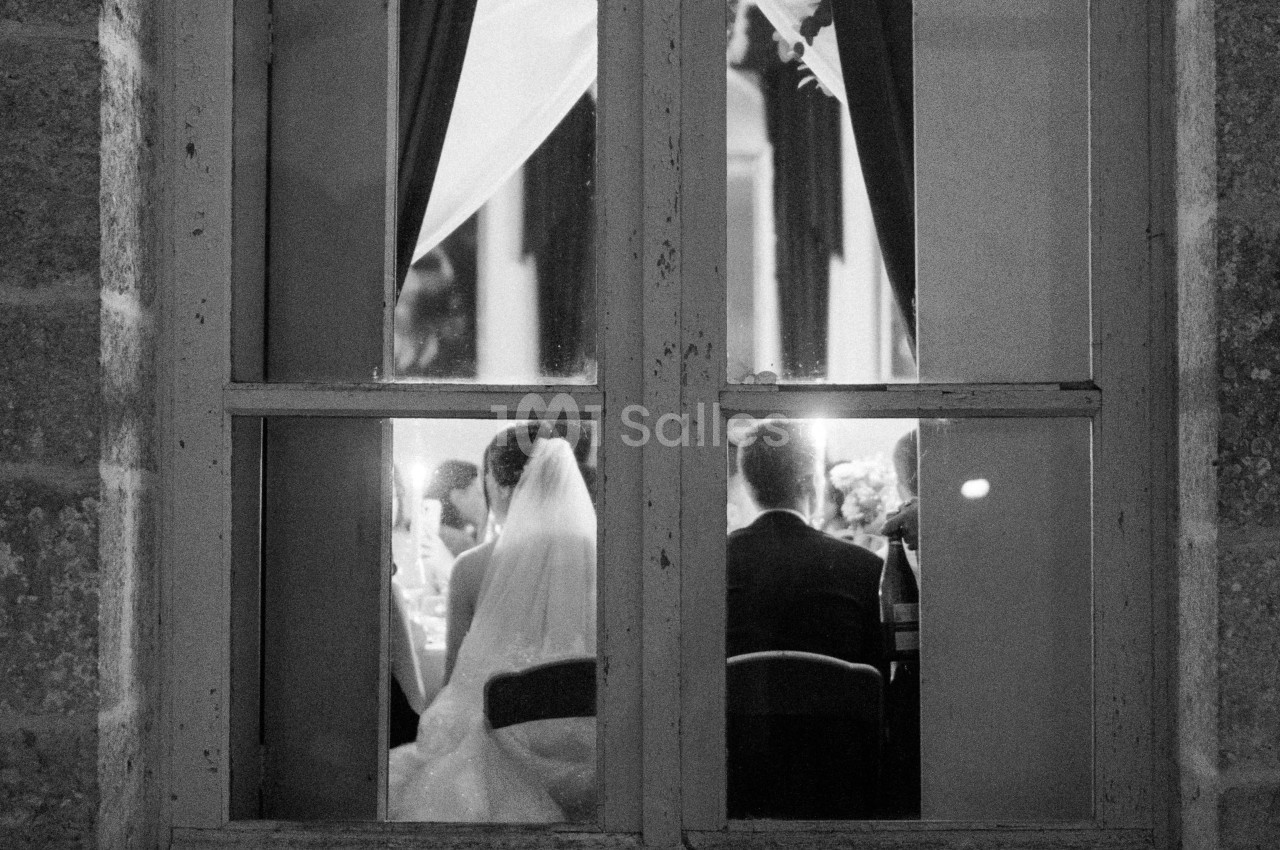 Vue à travers une fenêtre, un couple de mariés assis de dos lors d'une réception éclairée.