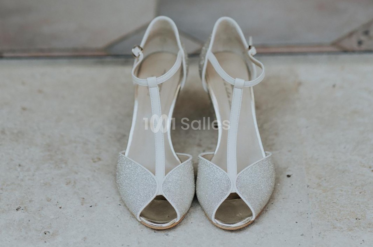 Chaussures à talons ouvertes en T, blanches et pailletées, posées sur un sol en pierre beige.