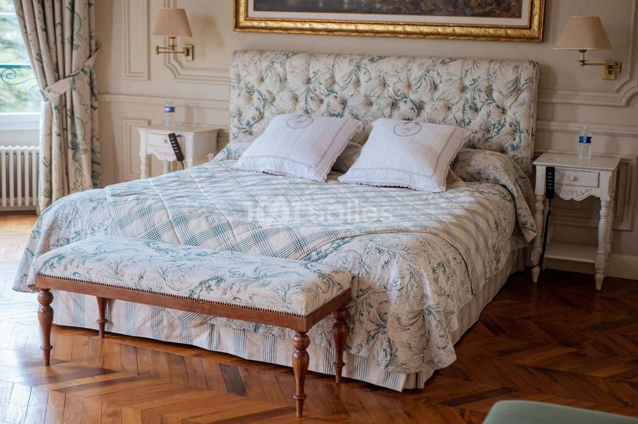 Lit double avec tête de lit capitonnée, banc en bois, tables de chevet et parquet en bois dans une chambre élégante.