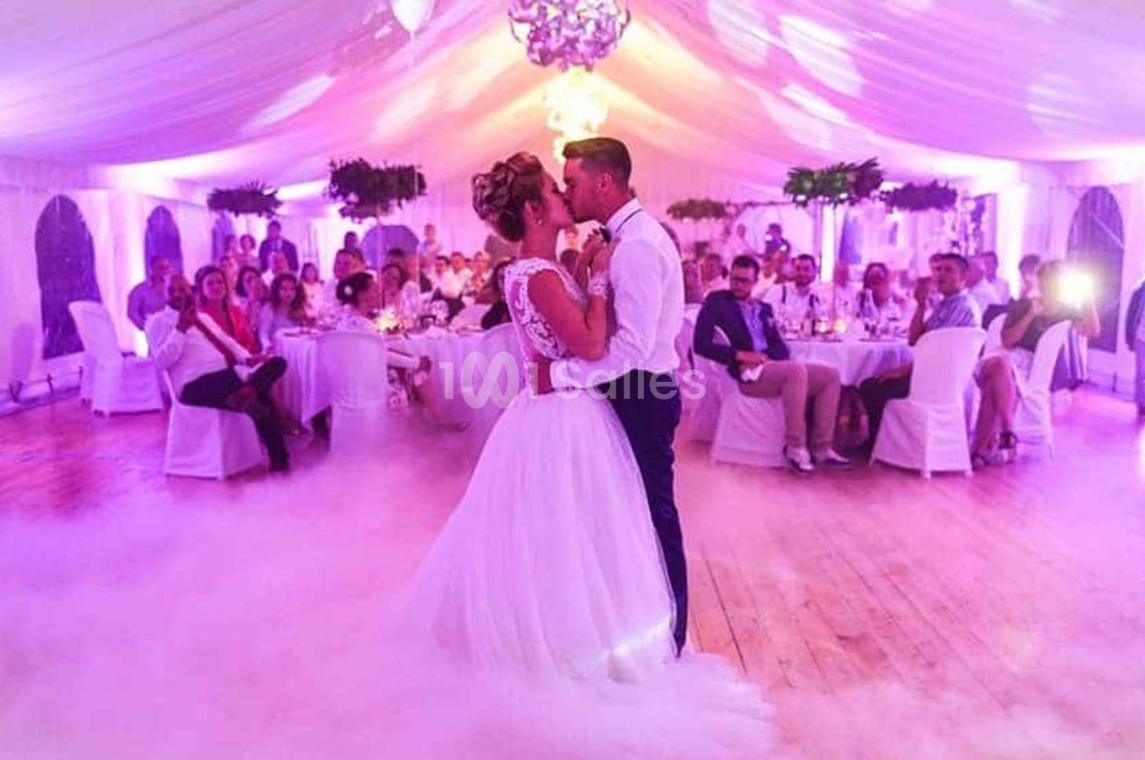 Un couple danse leur première danse de mariage dans une salle décorée avec un éclairage violet et un sol recouvert de fumée.