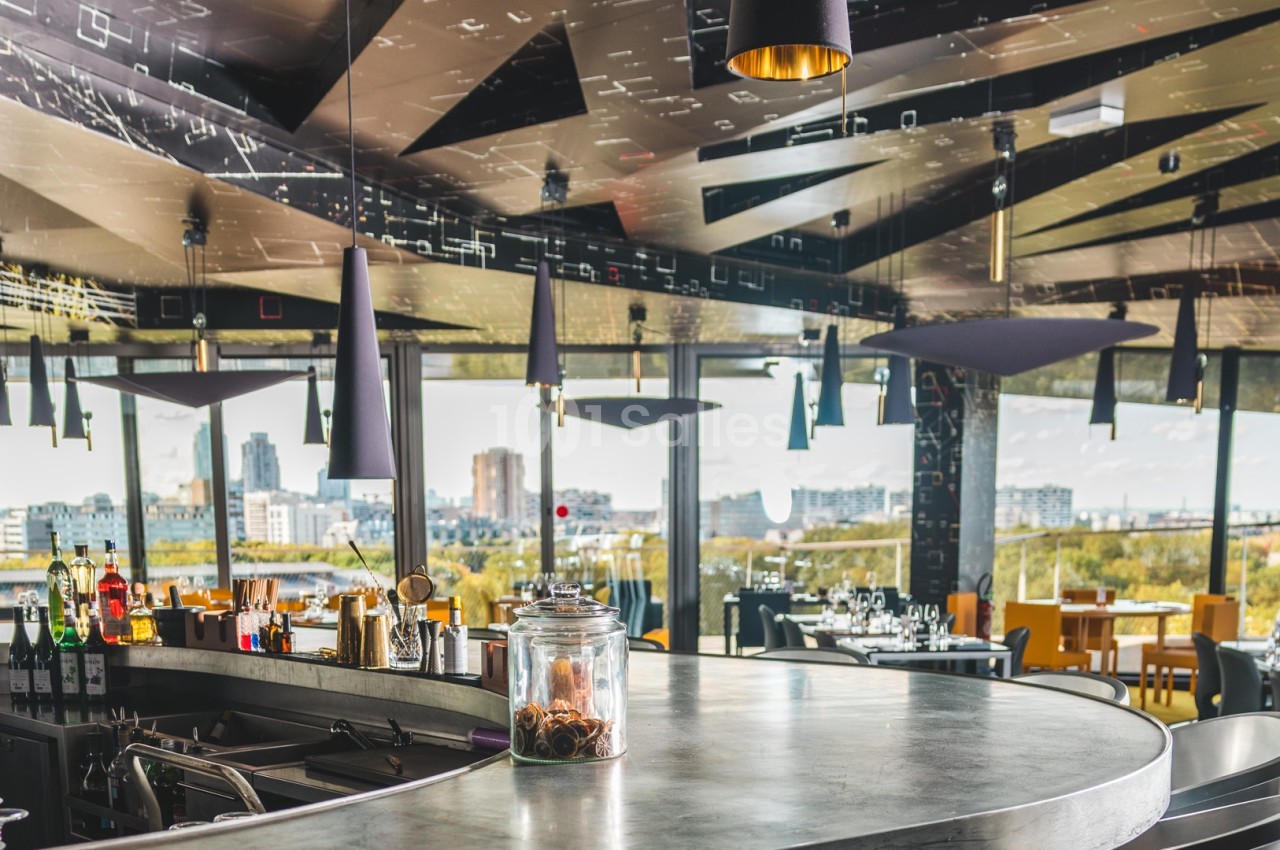 Intérieur d'un restaurant moderne avec un bar central, des tables dressées et une vue panoramique sur la ville.