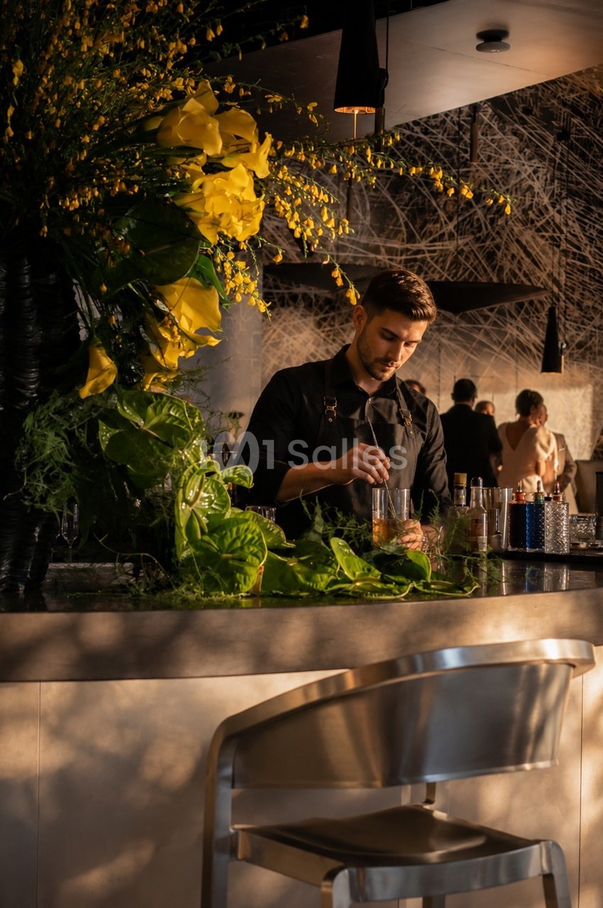Un barman prépare un cocktail dans un bar élégant, décoré de grandes compositions florales jaunes.