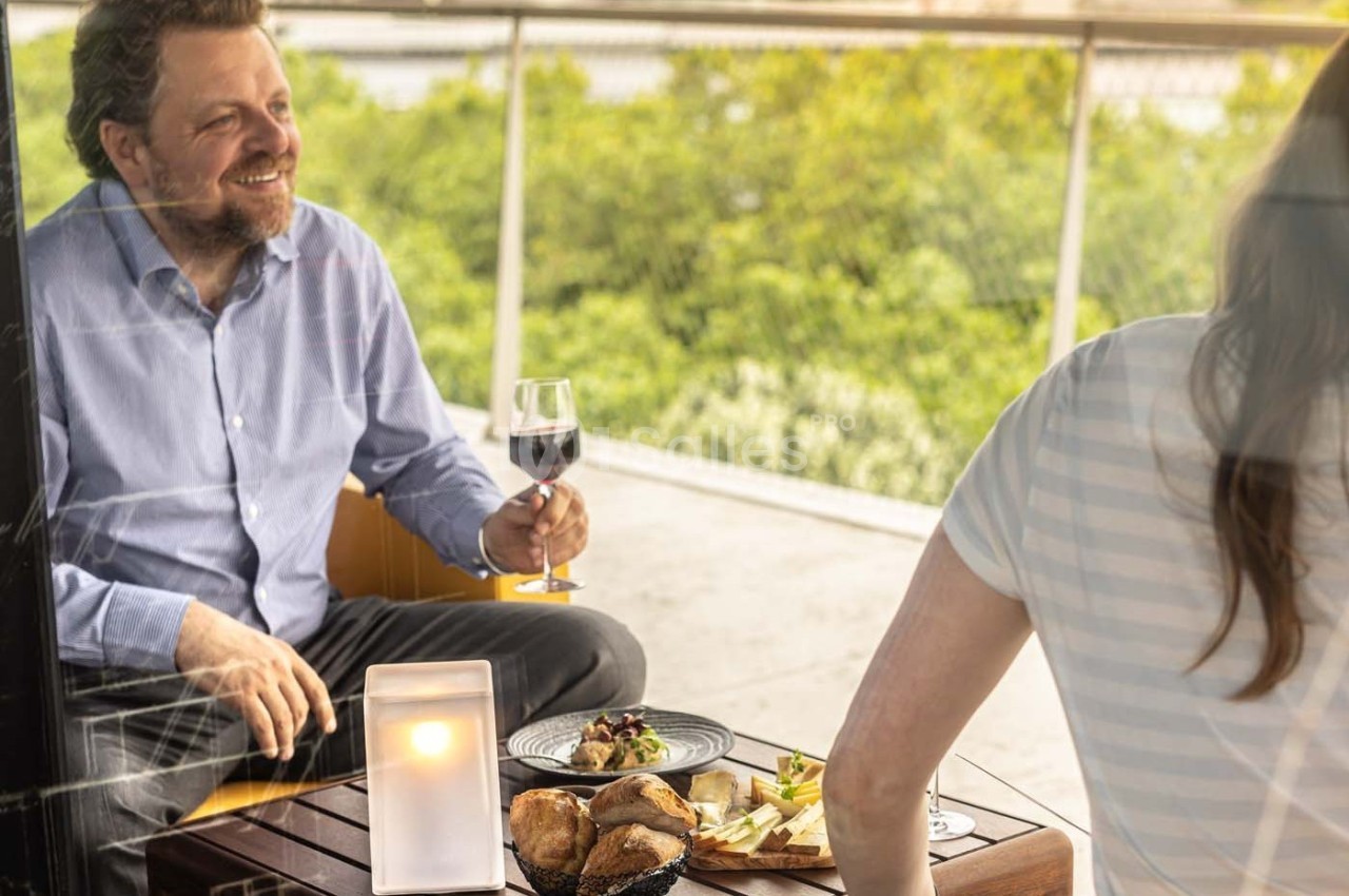 Un homme souriant tenant un verre de vin partage un repas en terrasse avec une femme, vue sur un paysage verdoyant.