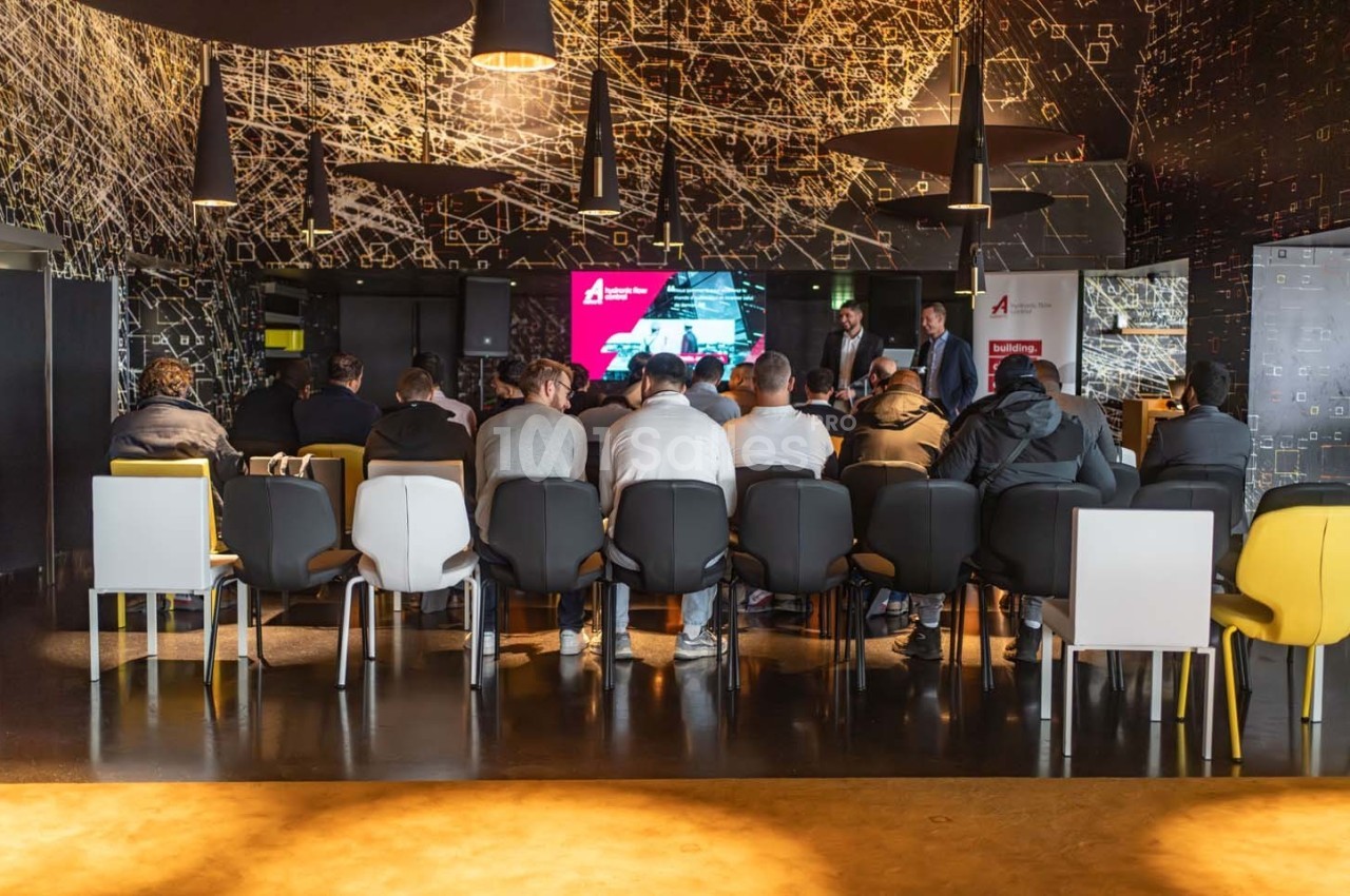 Groupe de personnes assises dans une salle de conférence moderne, écoutant une présentation sur un écran.