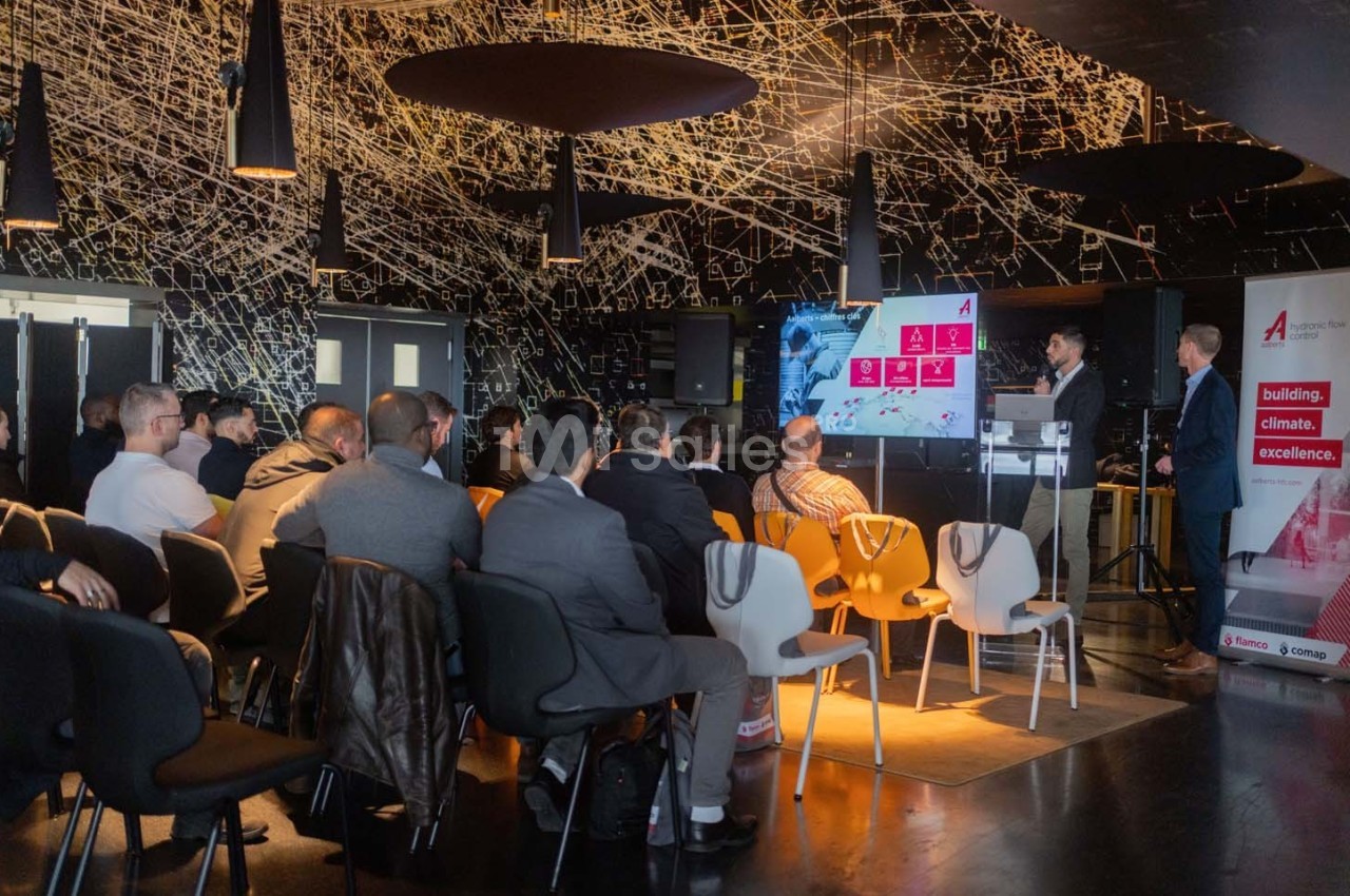 Un conférencier présente un diaporama à un public assis dans une salle moderne avec un plafond décoratif.
