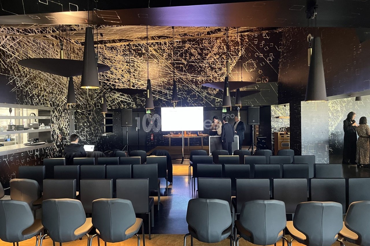 Salle de conférence moderne avec des rangées de chaises vides, un écran allumé et quelques personnes debout en arrière-plan.