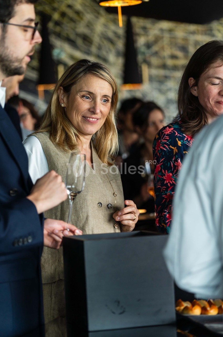 Une femme souriante tient un verre dans un cadre convivial avec d'autres personnes en arrière-plan.