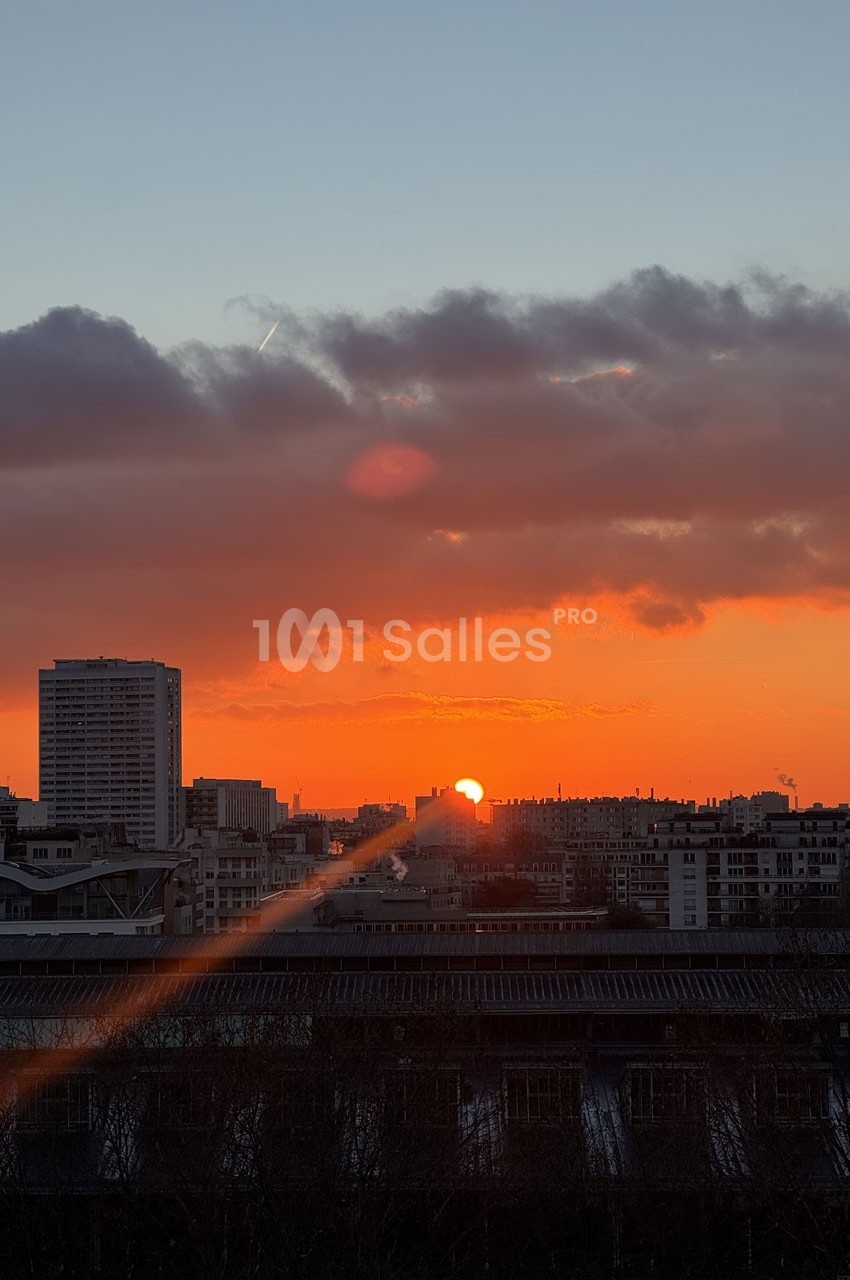Coucher de soleil sur une ville avec des immeubles, ciel orangé et nuages sombres à l'horizon.