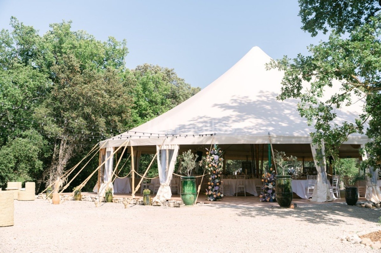 Tente blanche de style chapiteau installée en extérieur, entourée de végétation et décorée pour un événement.