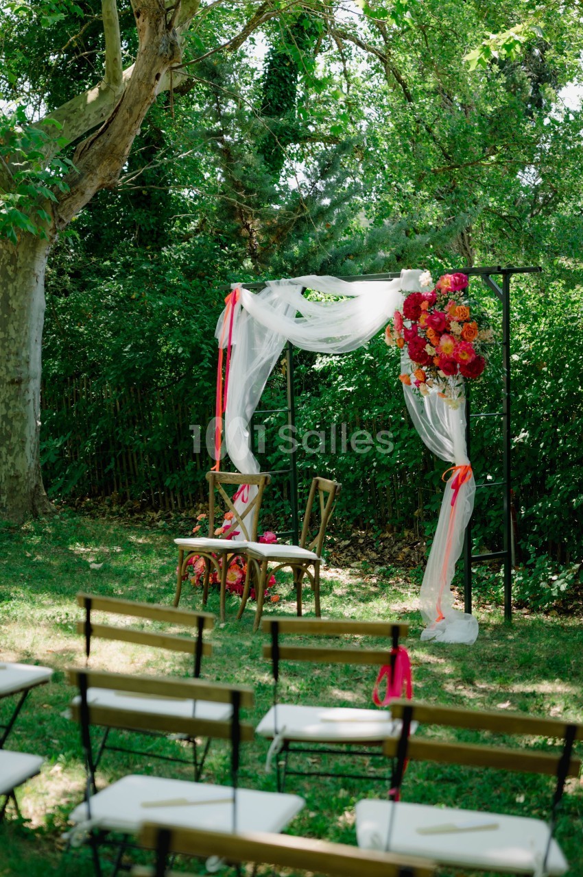 Arche de mariage décorée de fleurs rouges et blanches, entourée de chaises en bois dans un jardin verdoyant.