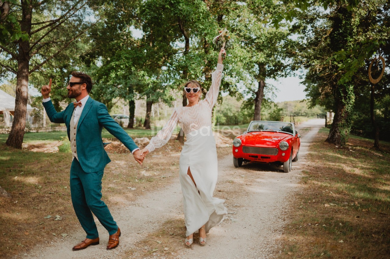 Un couple en tenue de mariage marche joyeusement sur un chemin bordé d'arbres, avec une voiture rouge en arrière-plan.